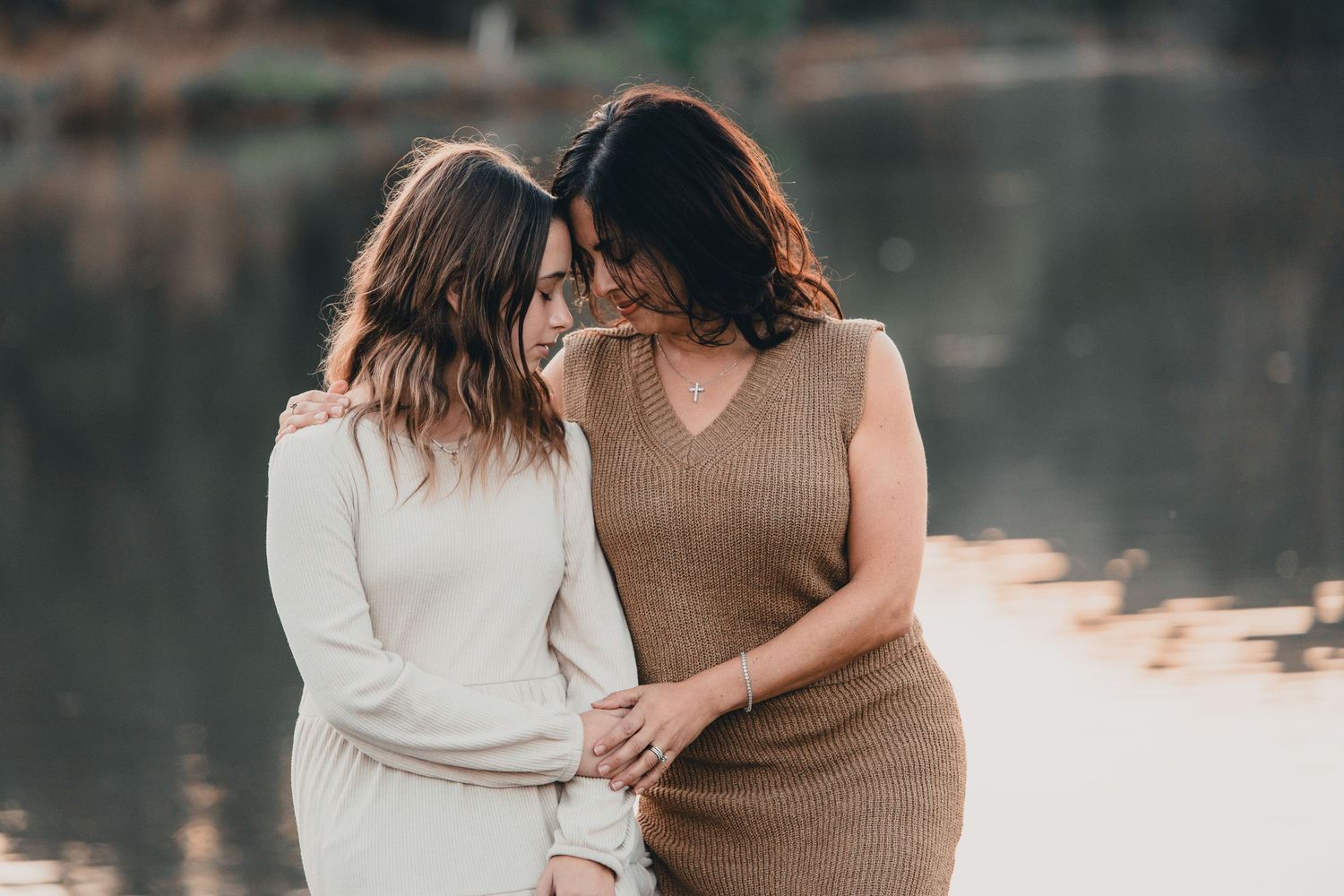 Two women embracing and smiling together near a reflective lake during golden hour.