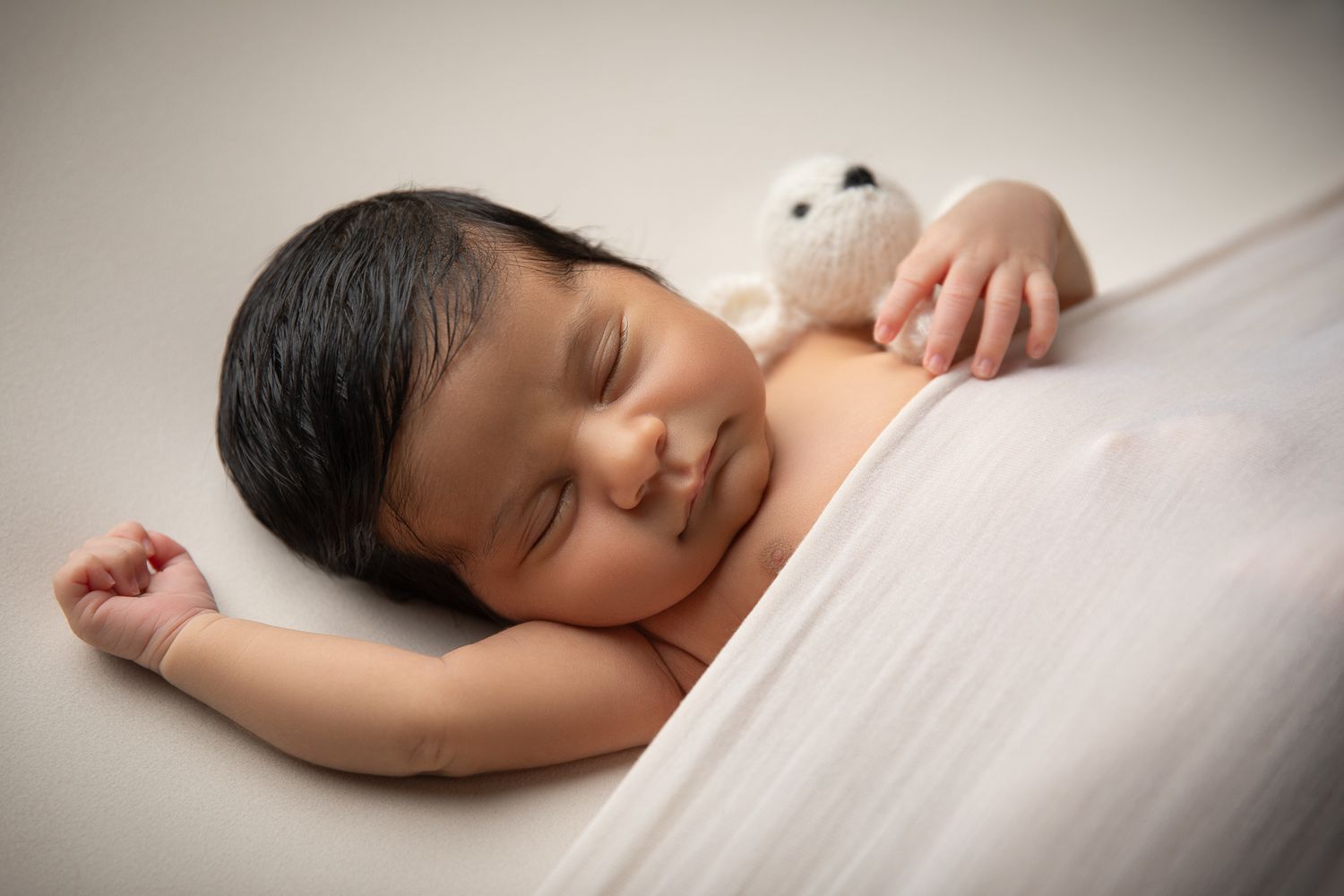 Sleeping newborn baby with dark hair lying beside a small white teddy bear on a soft surface.