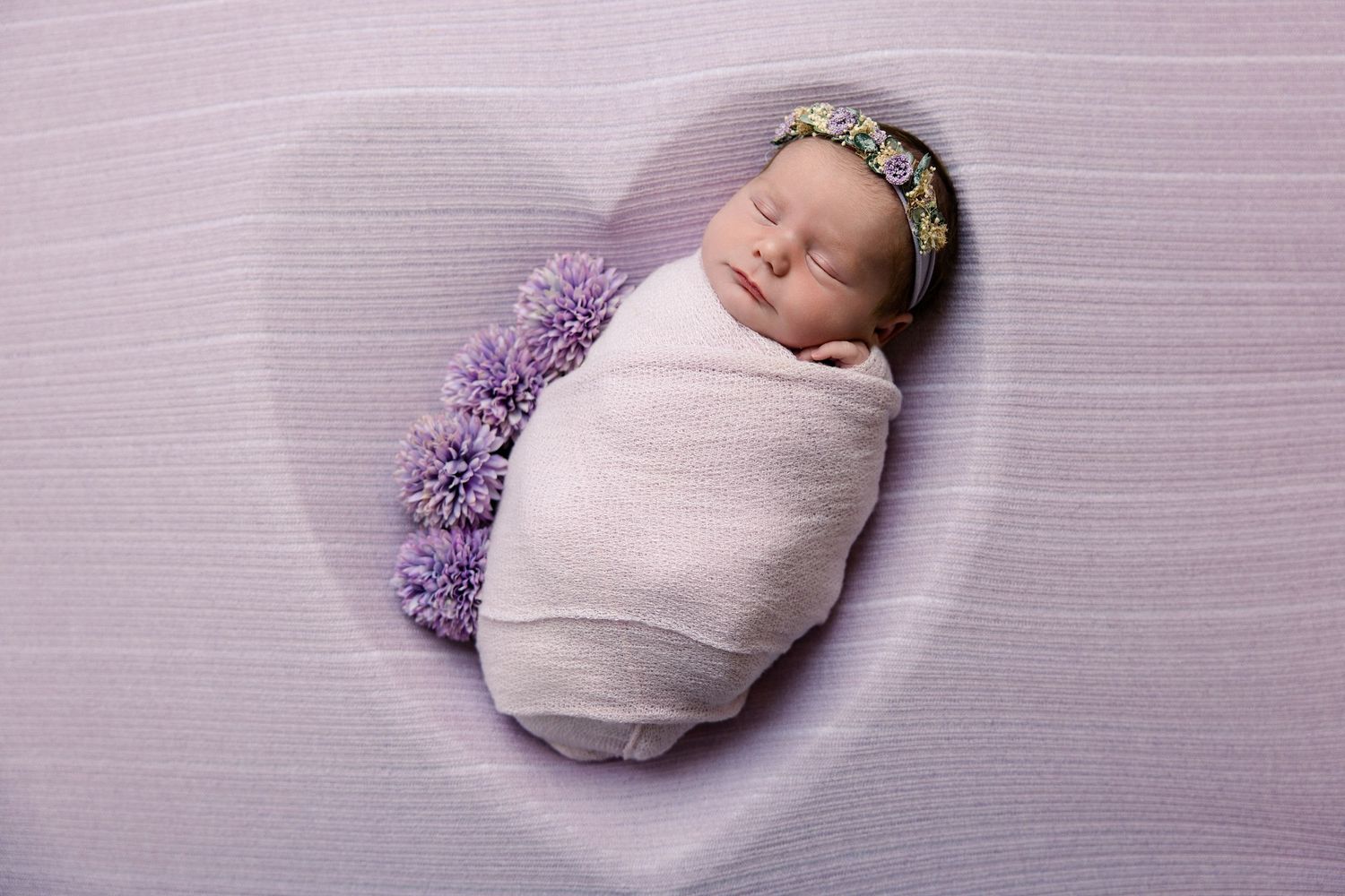 Newborn baby swaddled in white wrap with floral crown, sleeping in a heart-shaped purple backdrop.
