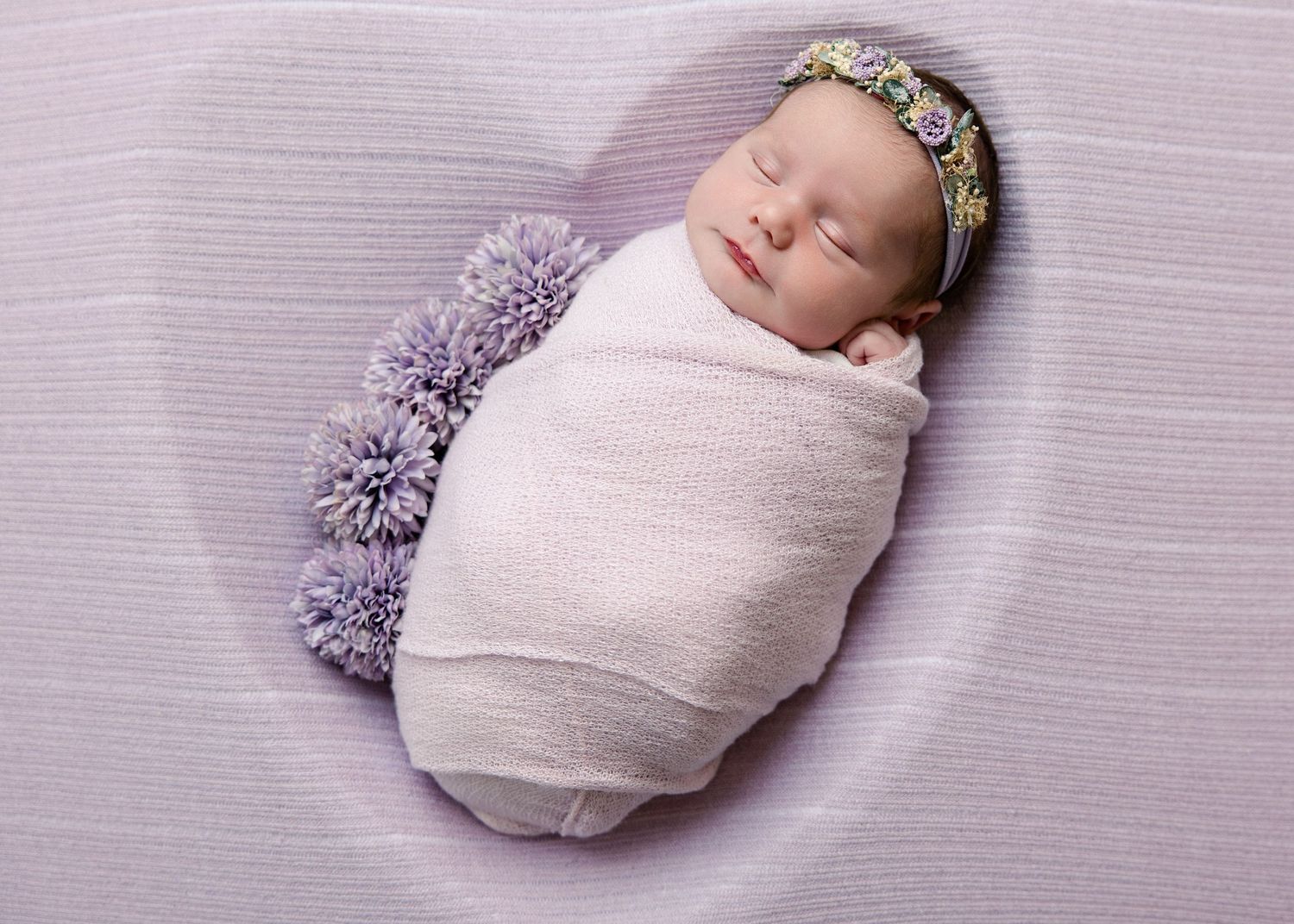 Newborn baby swaddled in white wrap with purple pom-poms, wearing floral headband, sleeping peacefully in heart-shaped bowl.