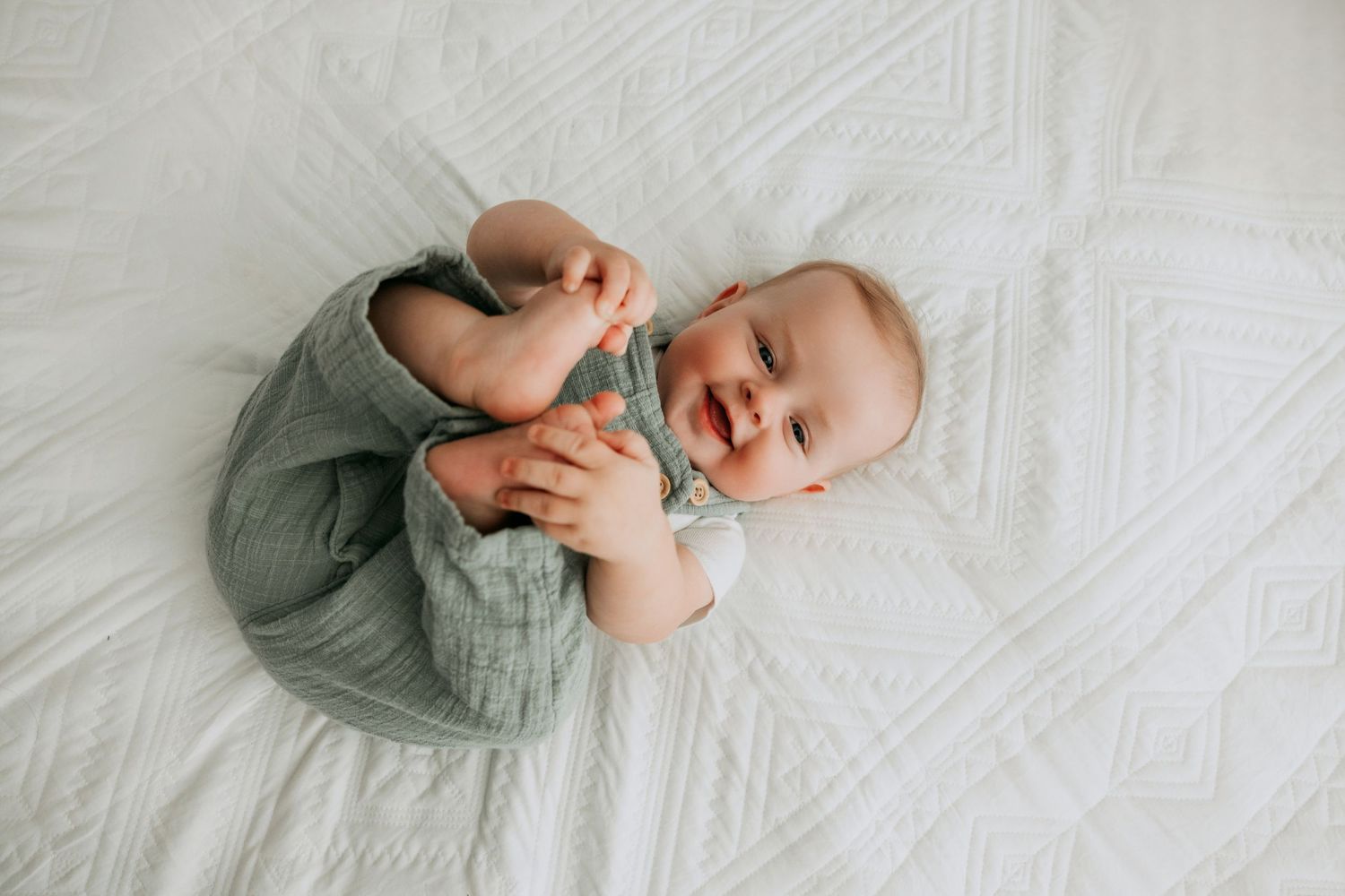 Smiling baby in green romper lying on white textured blanket, holding feet up in a joyful candid moment.