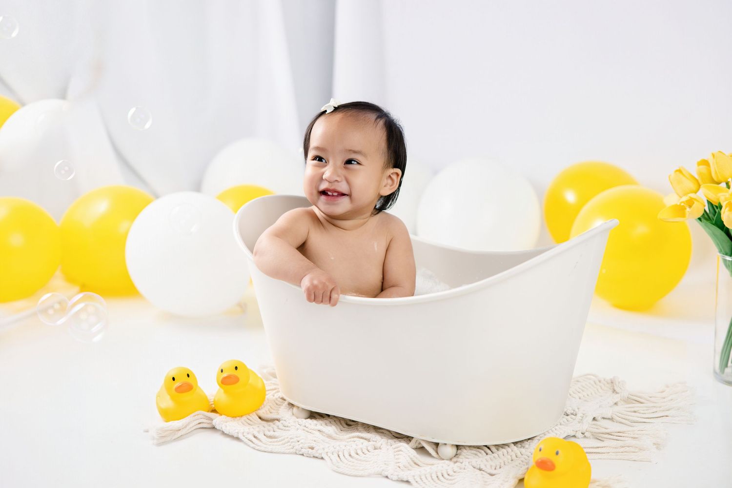 Smiling baby sitting in white bathtub surrounded by yellow and white balloons and rubber ducks.