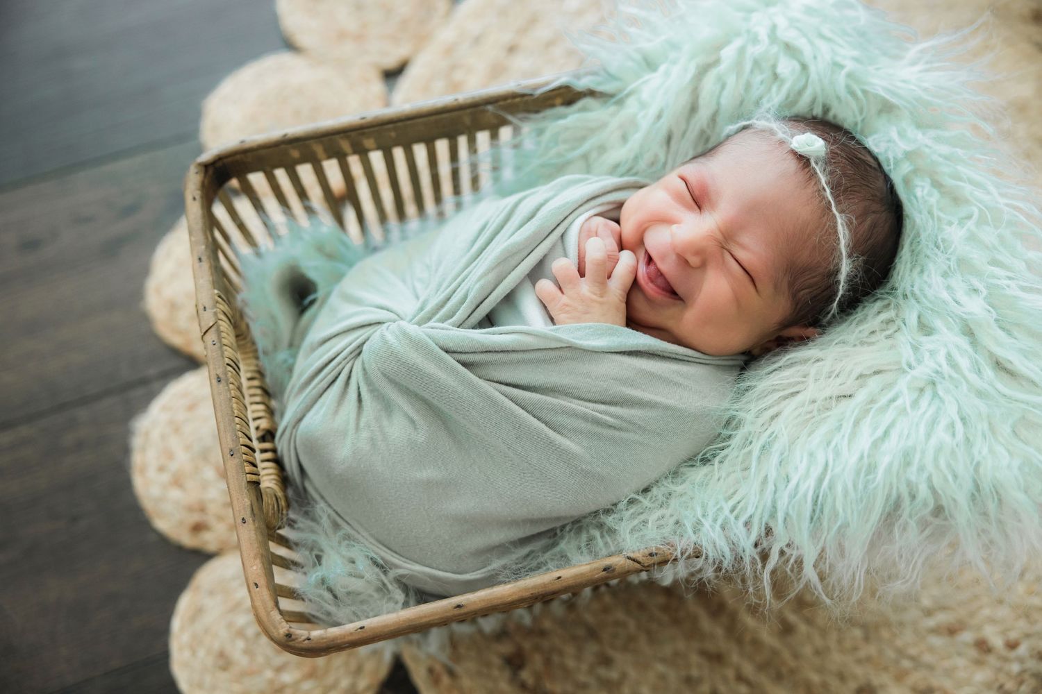 Smiling newborn baby wrapped in a mint green swaddle, resting in a wicker basket with fluffy teal fur.