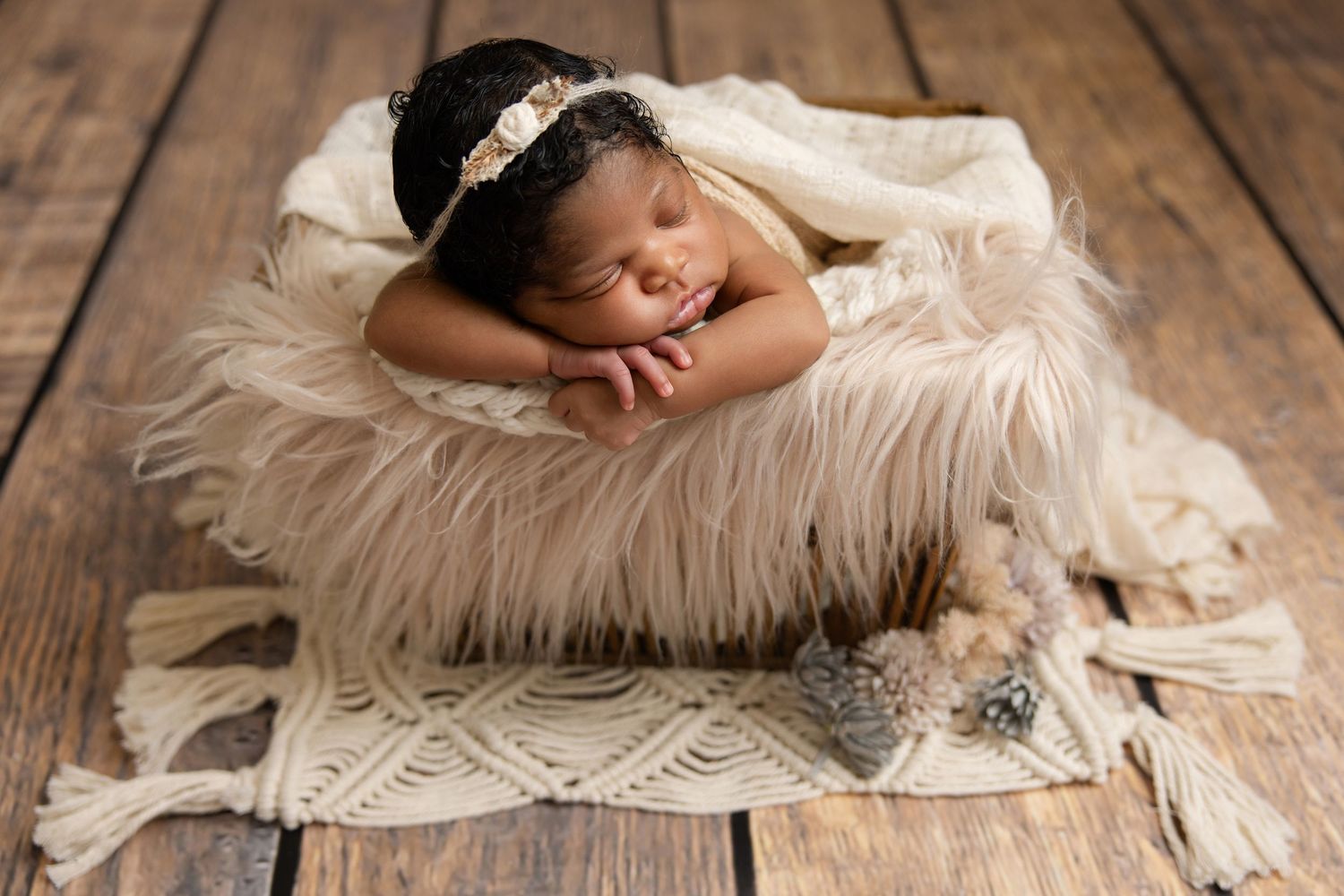 Newborn baby sleeping peacefully in a fluffy cream fur bowl with a delicate headband on a rustic wooden floor.