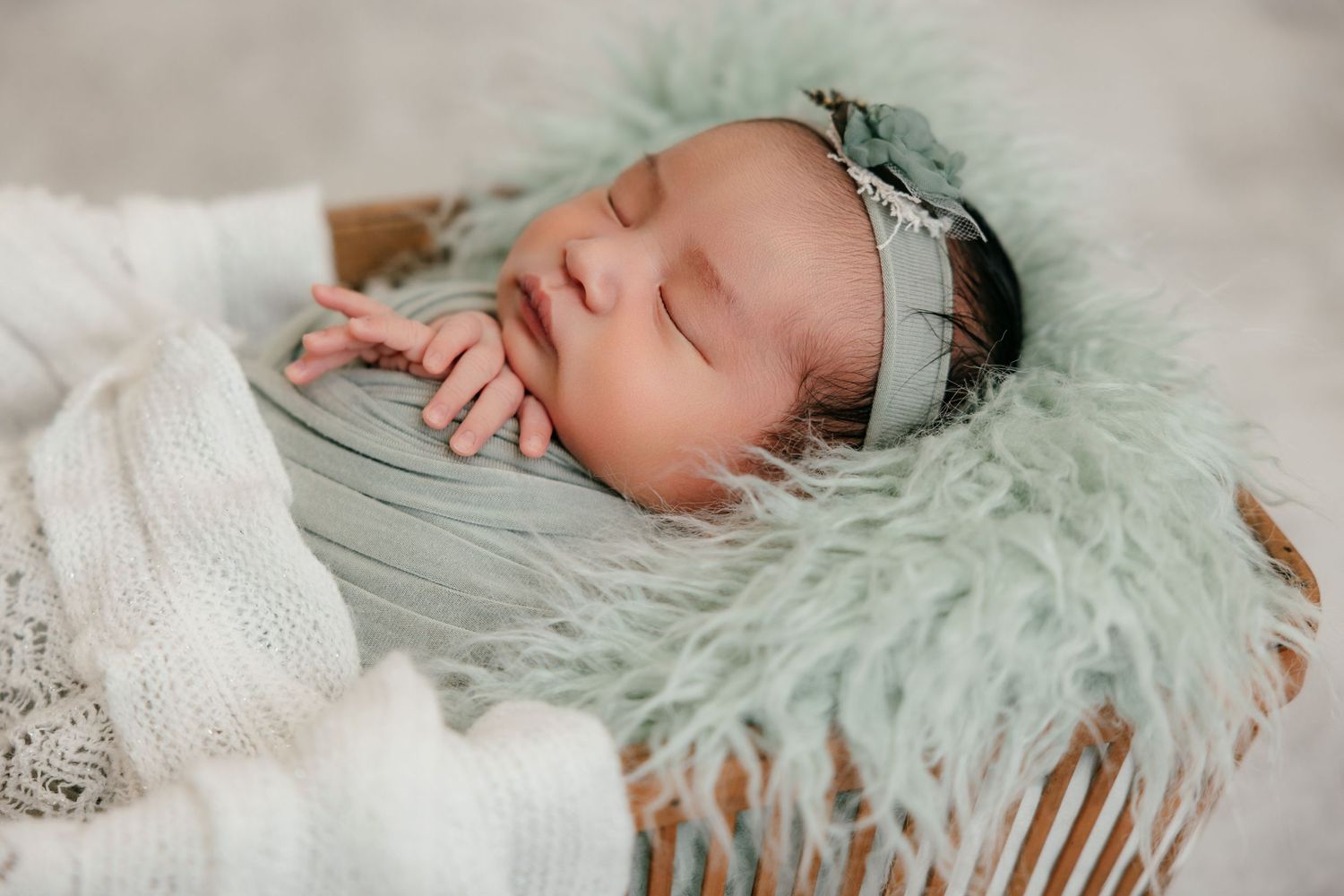Newborn baby sleeping peacefully in a basket with mint green fur, wrapped in a soft gray blanket and floral headband.