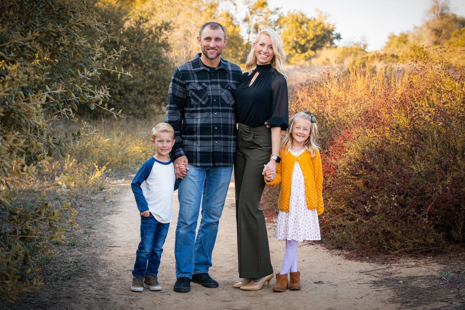 Family of four poses on a dirt trail surrounded by fall foliage in casual attire.