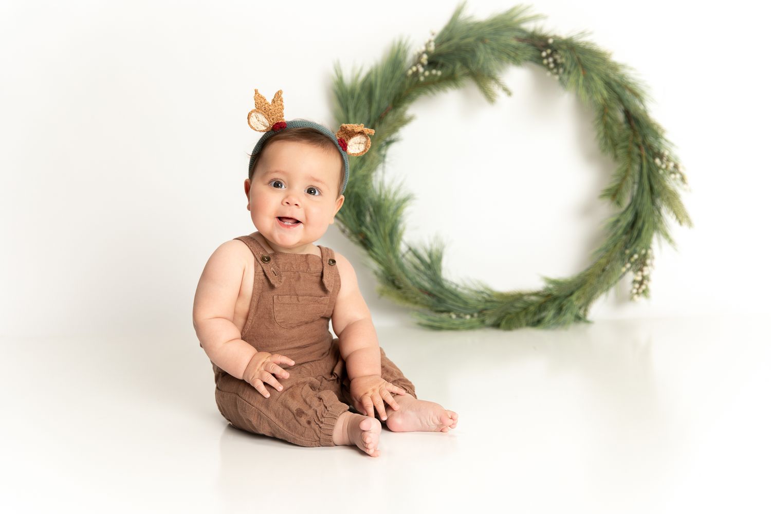 A smiling baby in brown overalls sits on white floor beside a green Christmas wreath with reindeer ear headband.