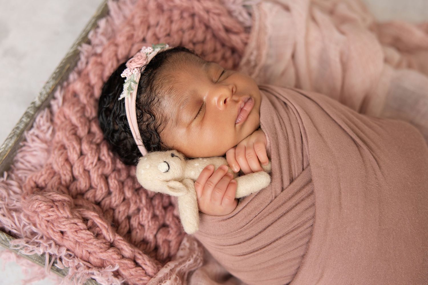 Sleeping newborn baby in pink swaddle clutching small bunny toy with floral headband in cozy wooden crate. Vista Newborn Photographer