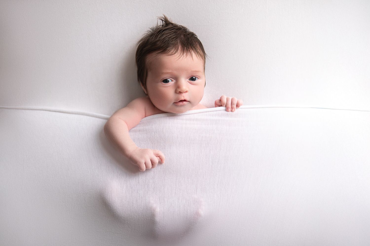 Newborn baby with dark hair peeks over a soft white blanket, lying on a white background in a serene studio photo.