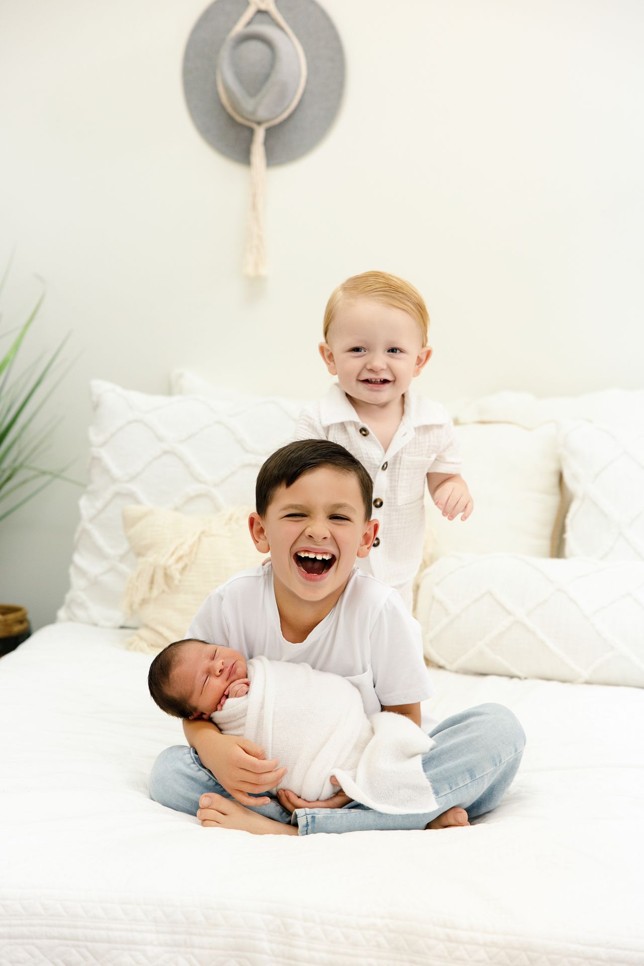 Two young boys joyfully pose on a white bed, one holding a sleeping newborn baby wrapped in a white swaddle.