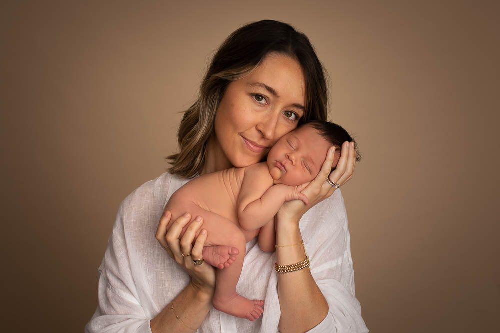 A smiling mother in a white shirt holds her sleeping newborn baby against a warm beige studio background.