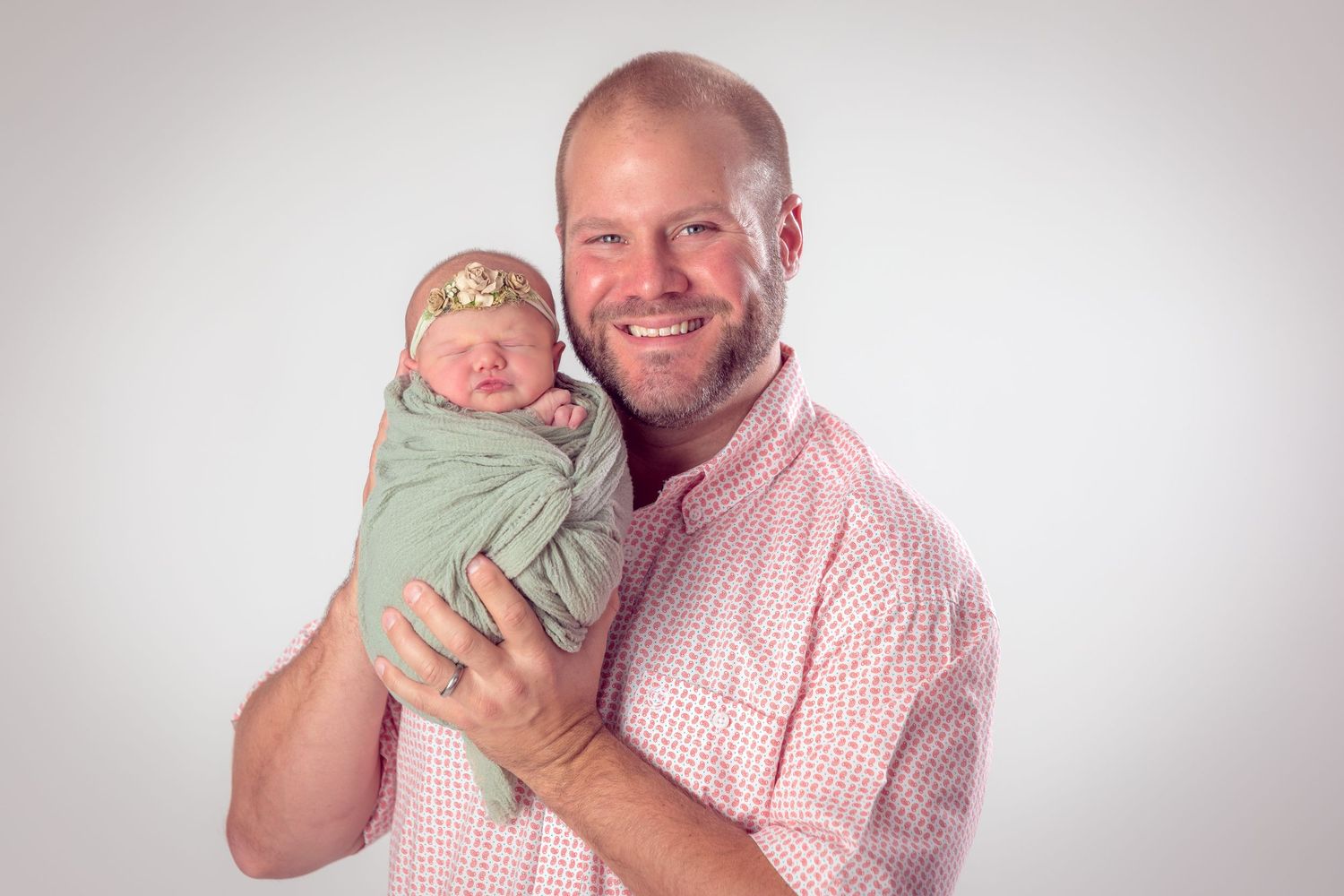 Smiling bald man in pink shirt proudly holds swaddled newborn baby wrapped in green with floral headband.
