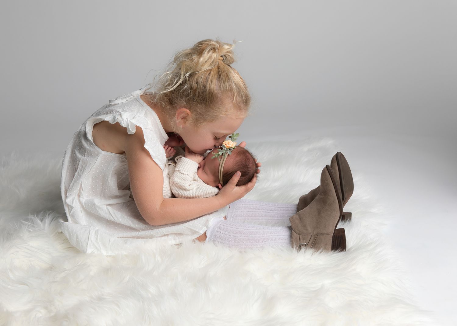 Young girl in white dress kissing newborn baby sibling on fluffy white rug in professional studio photo.