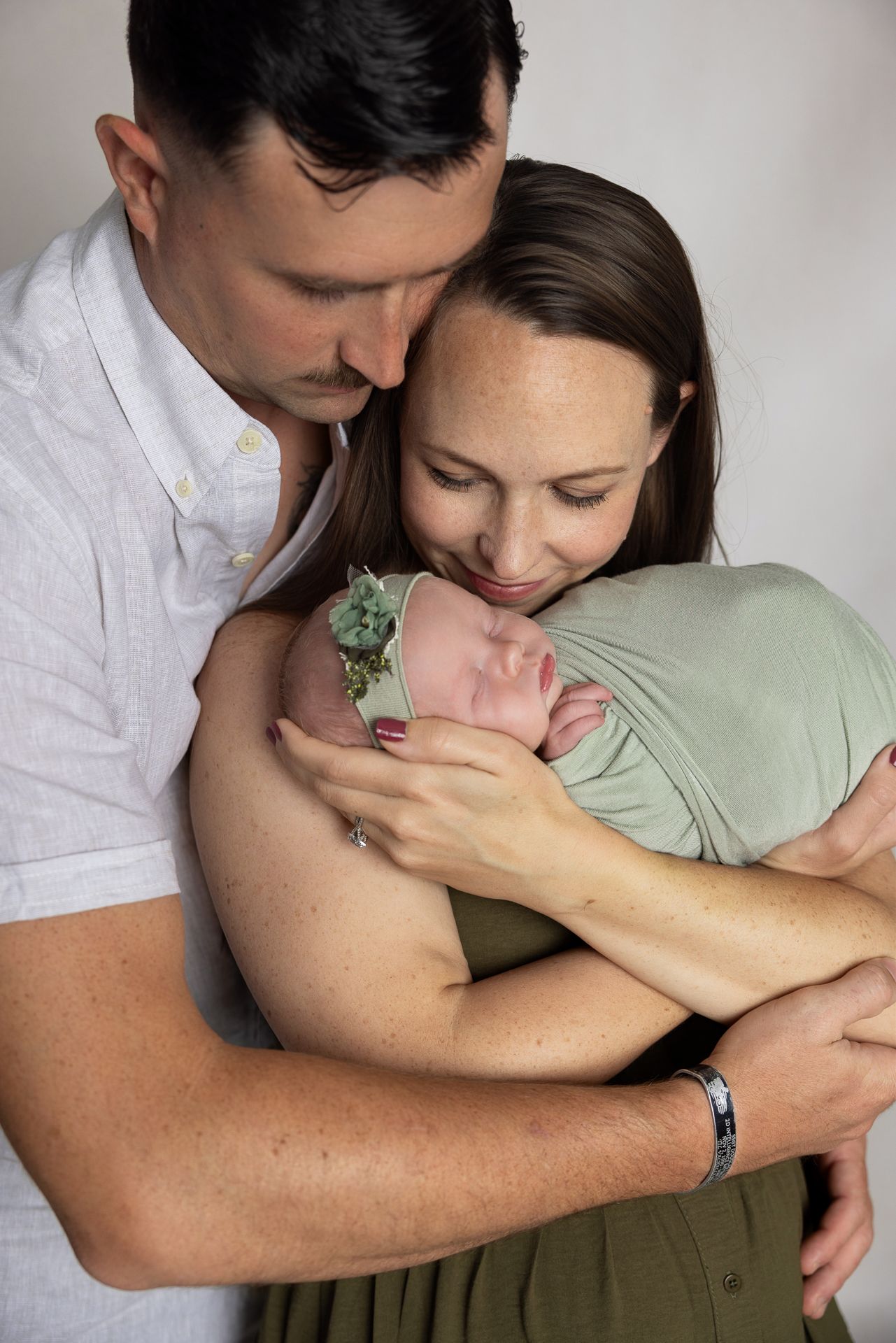 A couple tenderly cradles their sleeping newborn baby wrapped in a green swaddle with a floral headband.