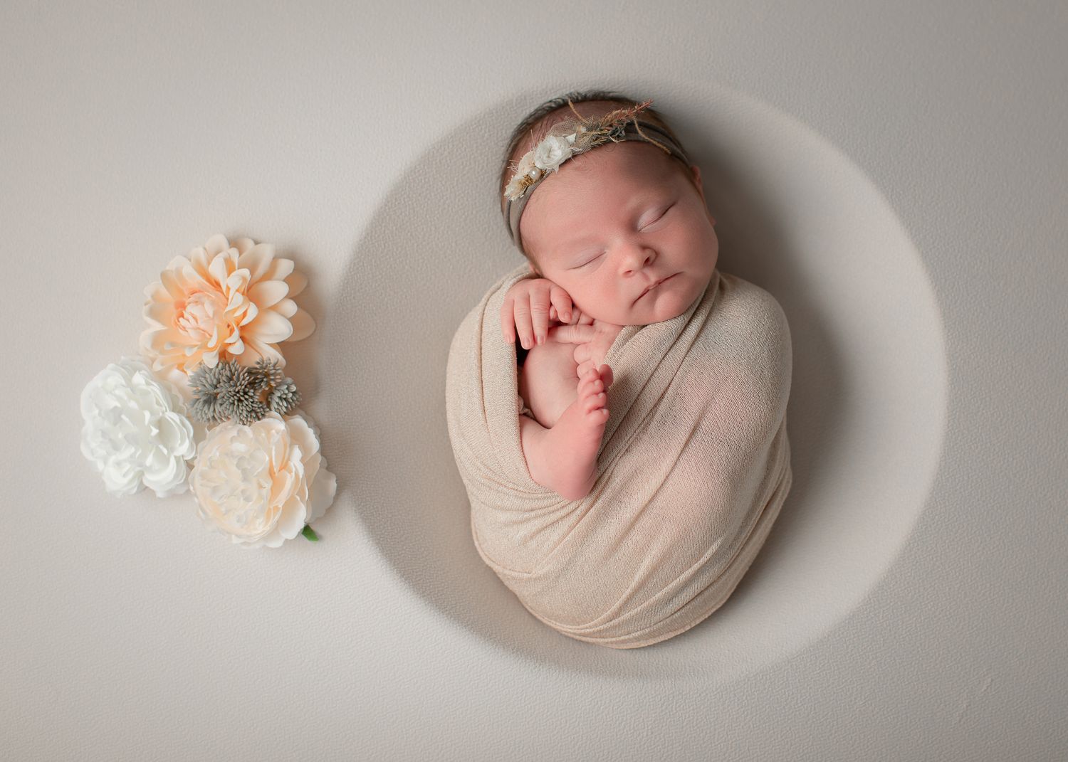 Sleeping newborn baby swaddled in cream wrap with floral headband, posed in white bowl with peach and white flowers.