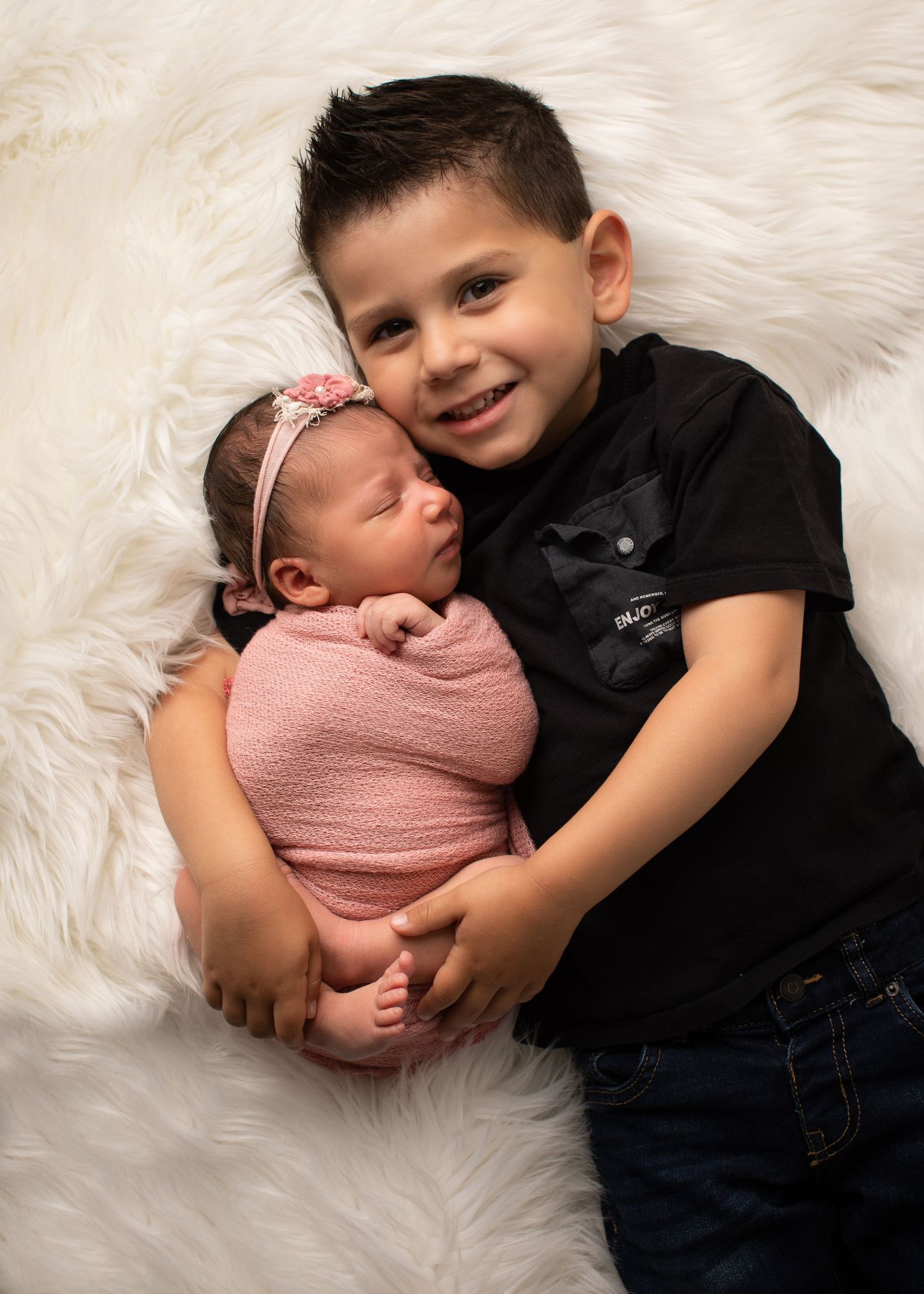 Smiling young boy in black shirt holds newborn baby girl wrapped in pink on a white fluffy blanket.