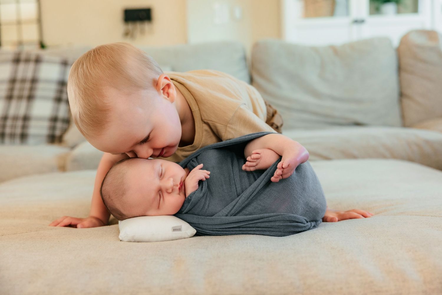 Toddler boy gently kissing sleeping newborn baby sibling wrapped in gray swaddle on a couch.