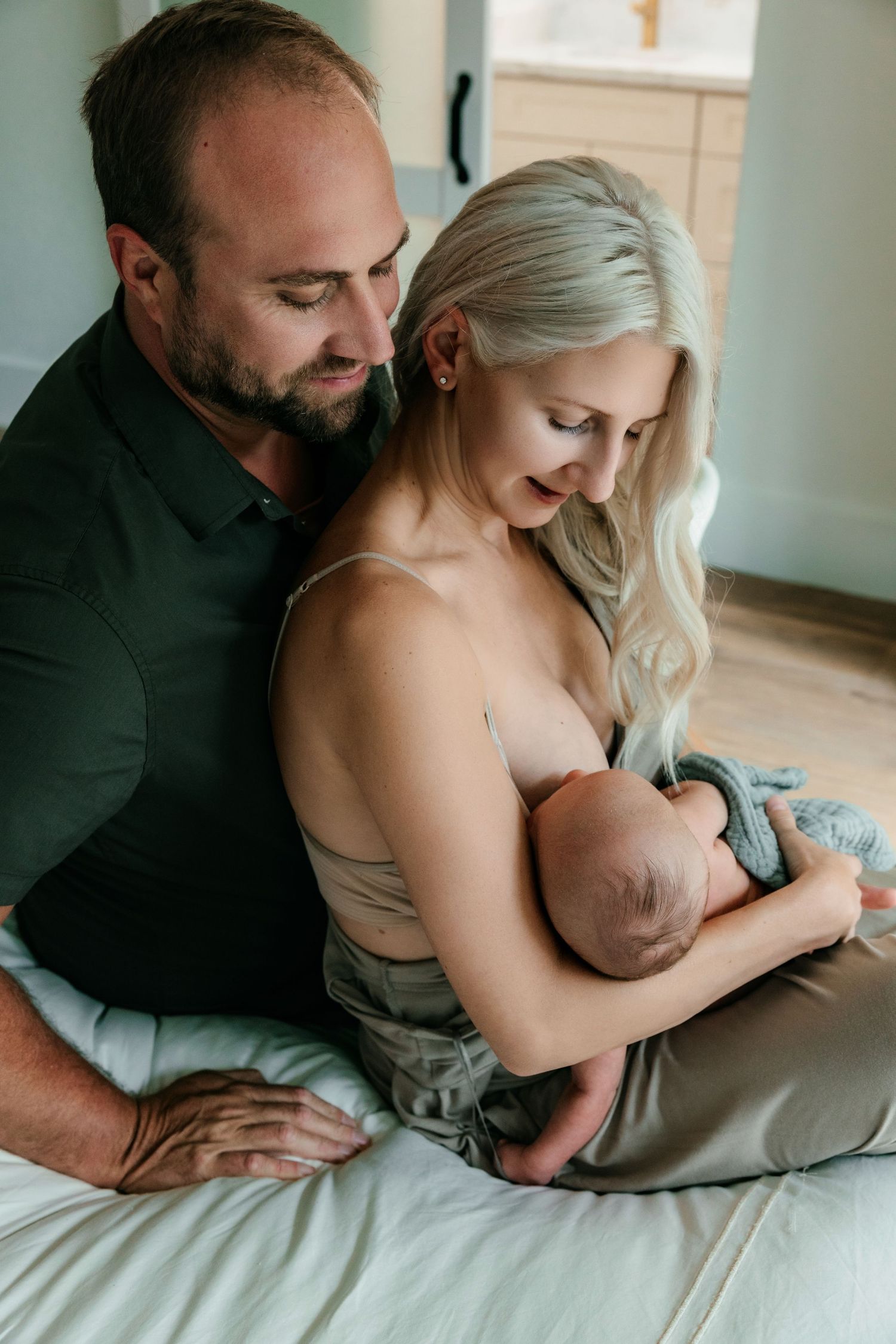 A couple sits on a bed, lovingly gazing at their newborn baby wrapped in a blue blanket.