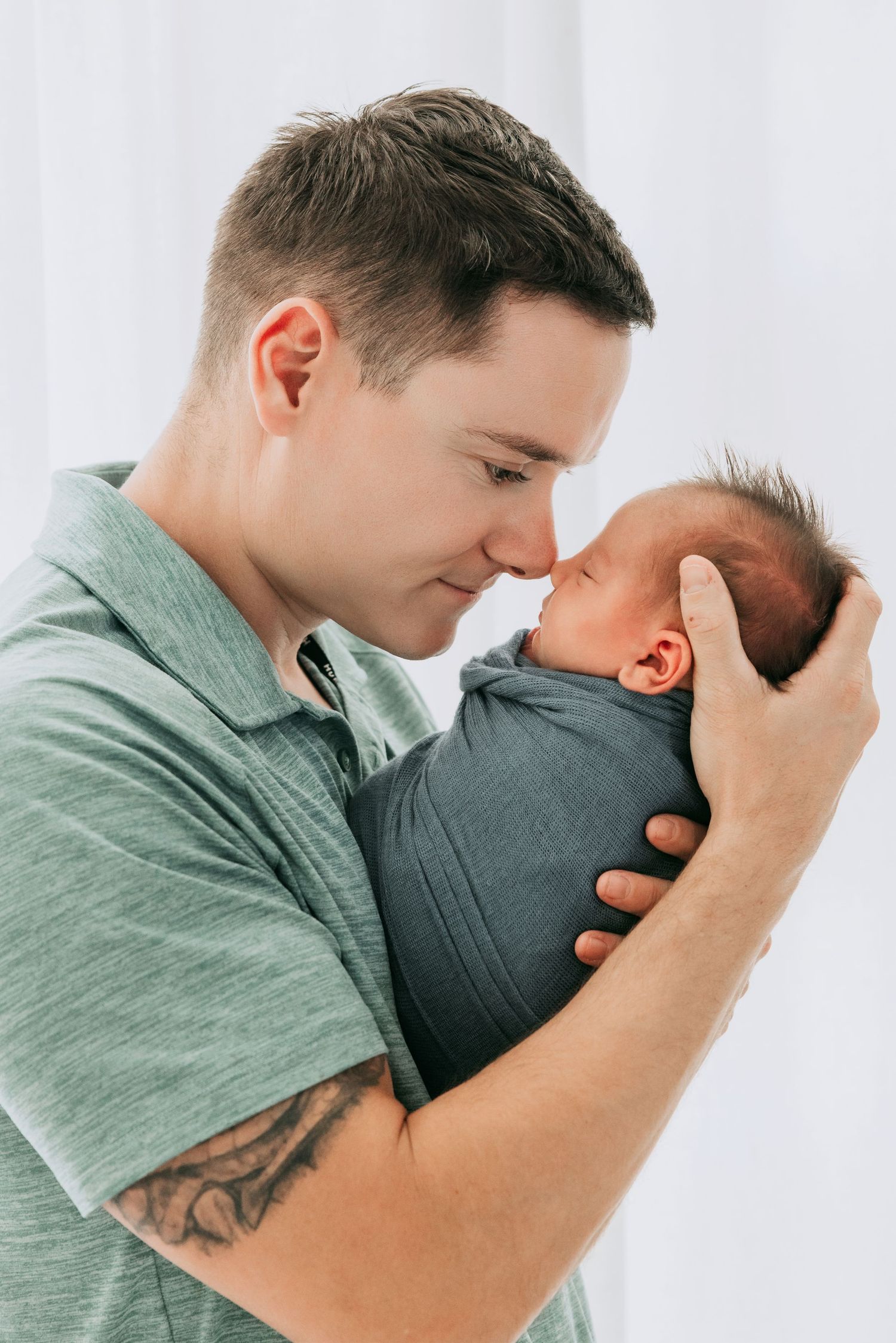 Father in green shirt tenderly touches noses with swaddled newborn baby, sharing a precious bonding moment.