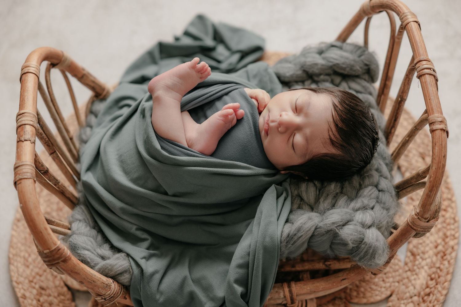 Newborn baby sleeping peacefully wrapped in a sage green swaddle blanket inside a wicker bassinet.