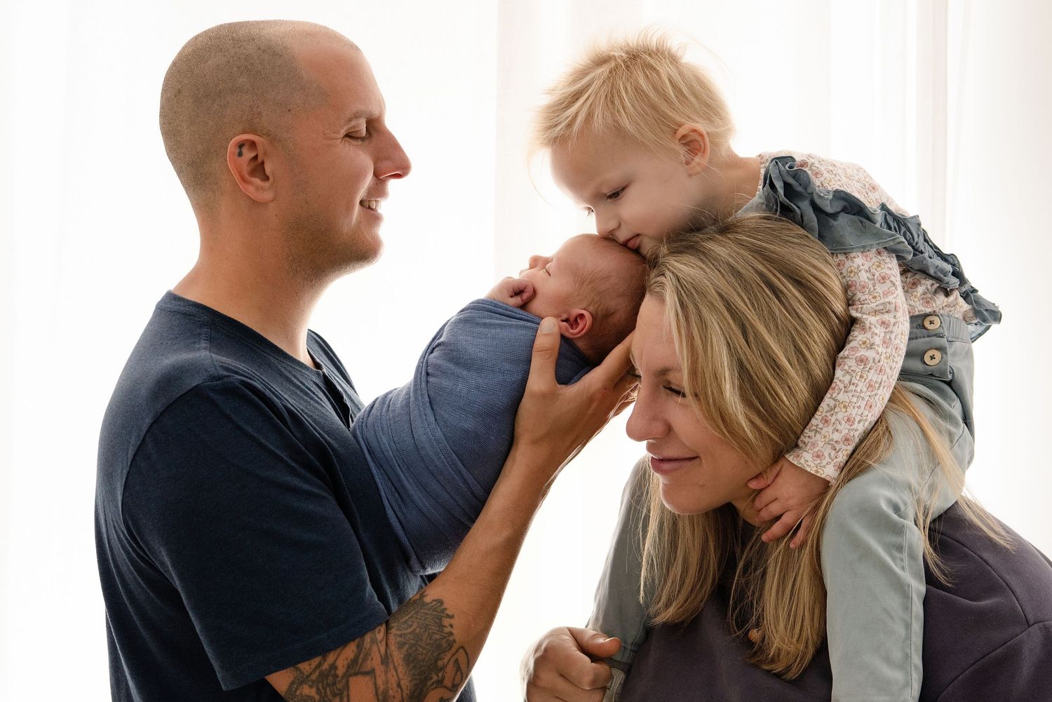 A tattooed father holds a newborn while a toddler kisses the baby atop the smiling mother's shoulders.