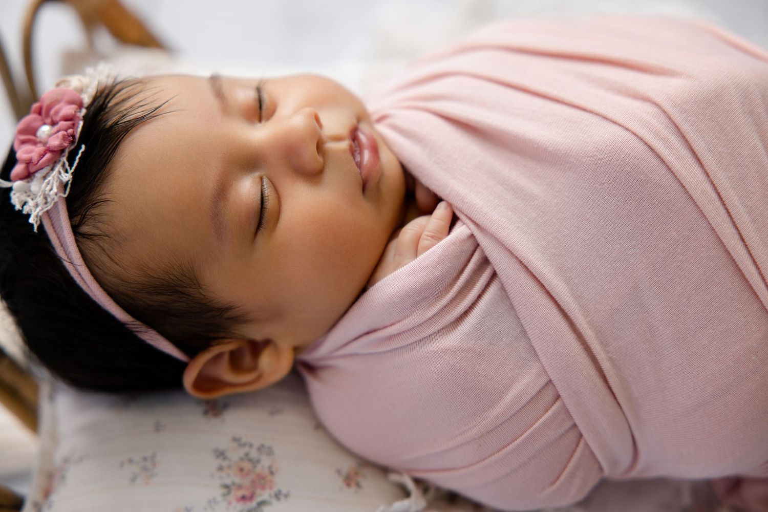 Sleeping newborn baby swaddled in a soft pink wrap, wearing a floral headband, peacefully resting.