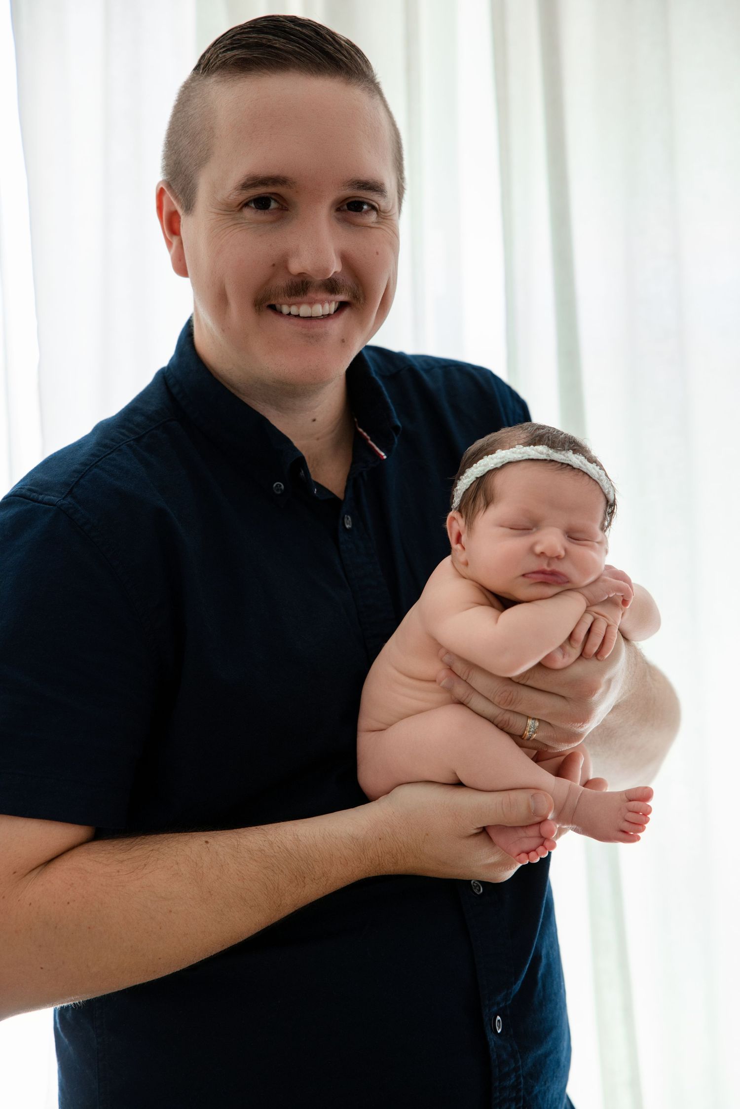 Smiling man in navy shirt holds sleeping newborn baby with white headband near bright window.