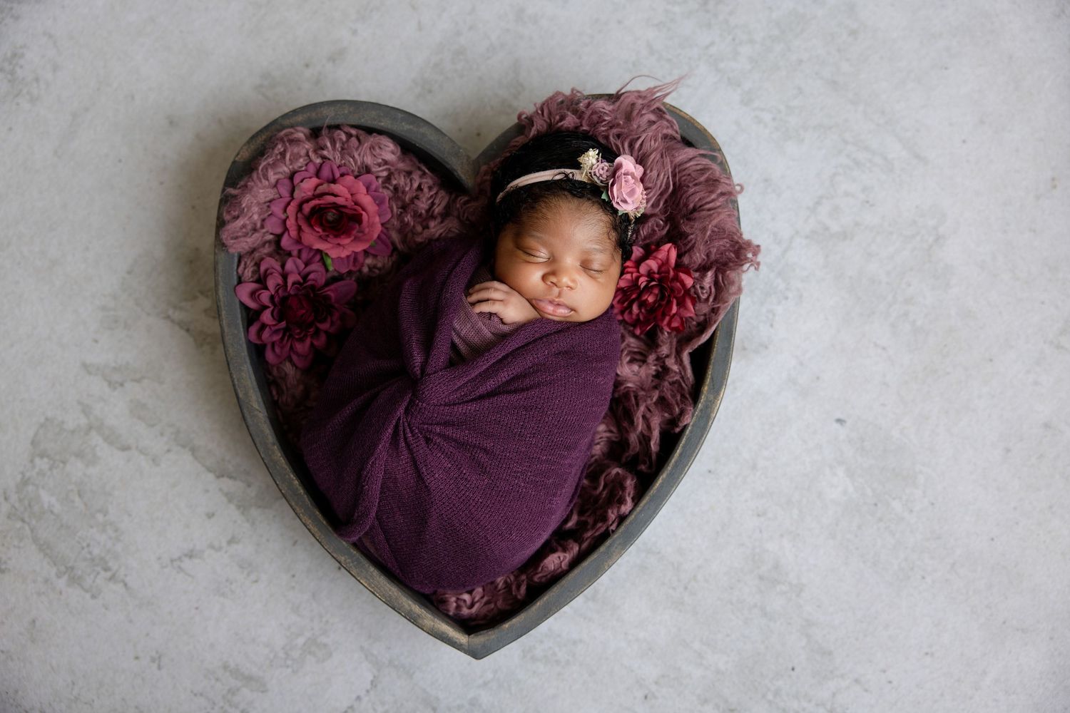 Sleeping newborn baby wrapped in purple swaddle in heart-shaped bowl with pink flowers.