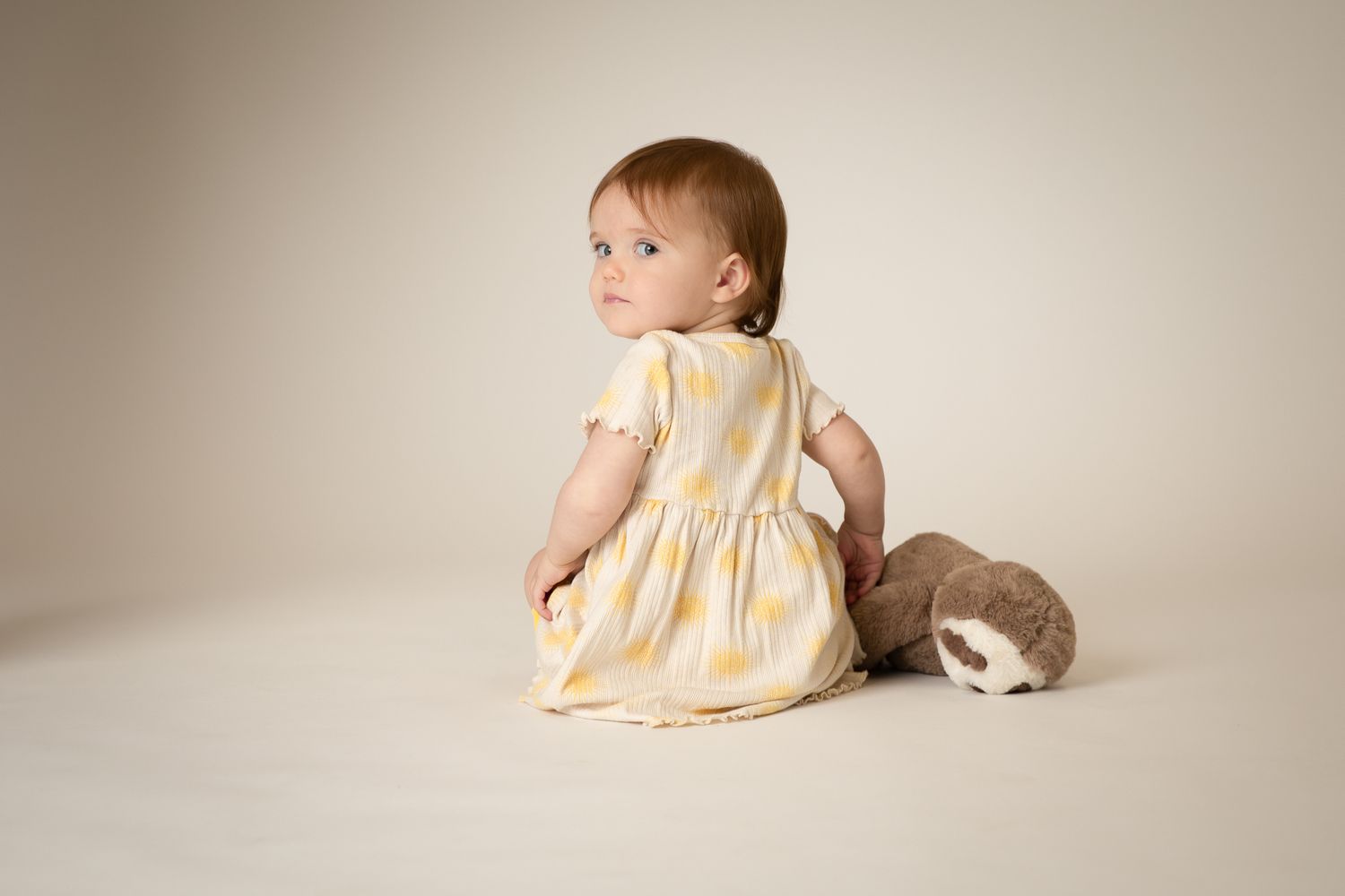 Baby girl in yellow striped dress sitting with plush sloth on neutral background.