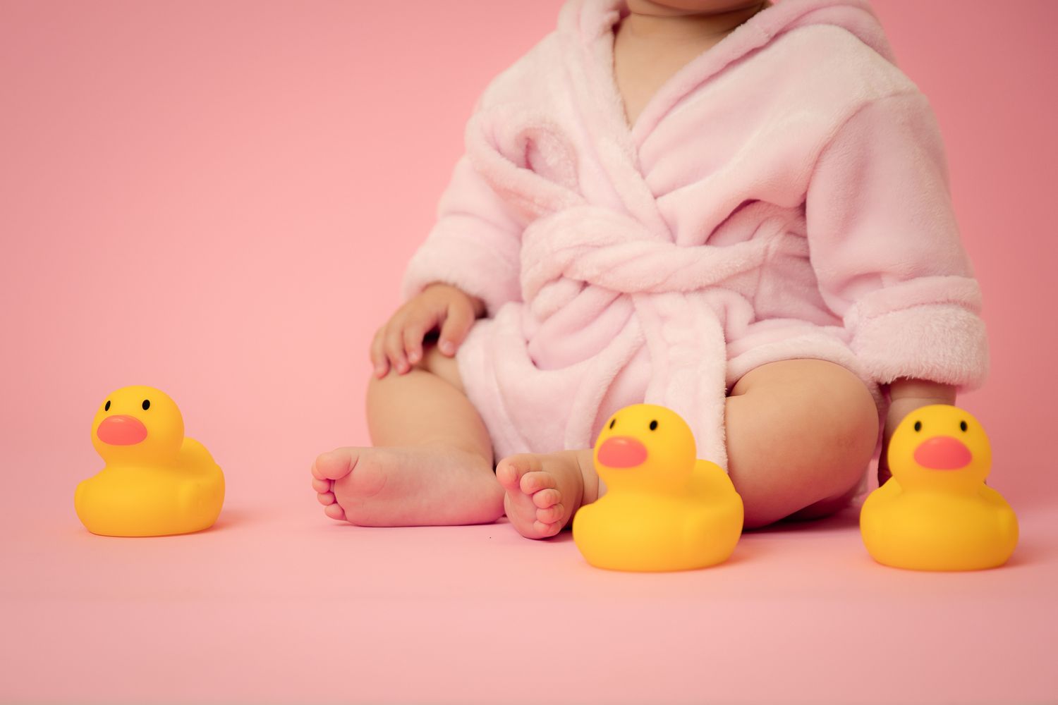 Baby in pink robe sitting with yellow rubber ducks on pink background.