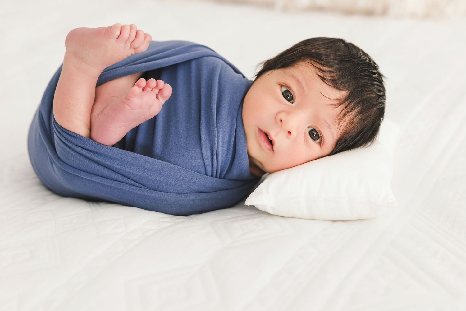 Alert newborn baby wrapped in blue swaddle blanket lying on white pillow during newborn photography session in Oceanside CA