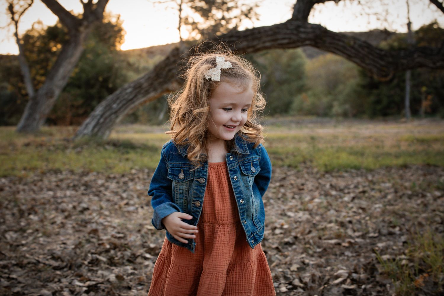 Curly-haired toddler girl in denim jacket and rust dress smiles in autumn outdoor setting.