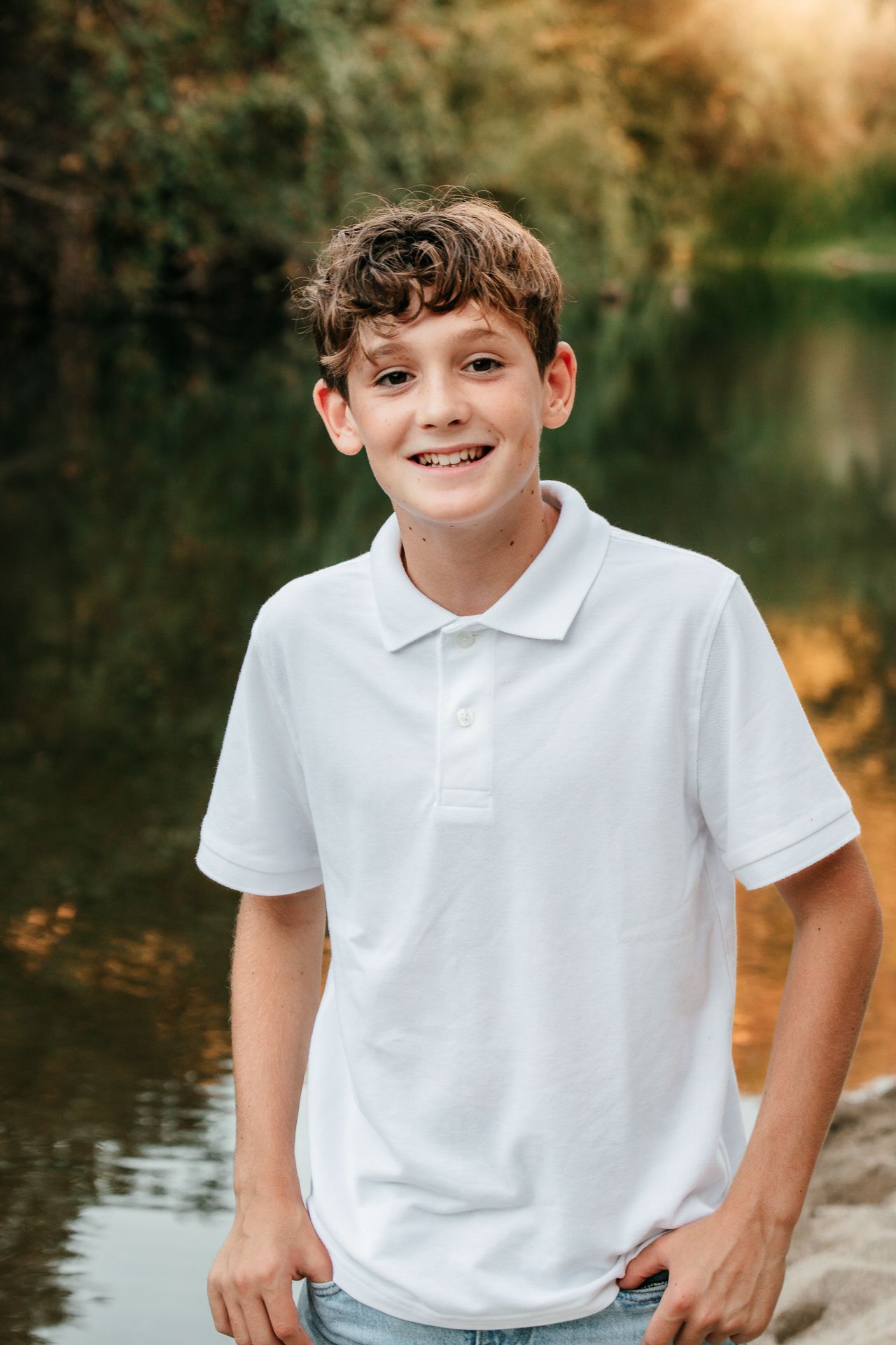 A smiling teenage boy in a white polo shirt stands near a reflective pond surrounded by trees.