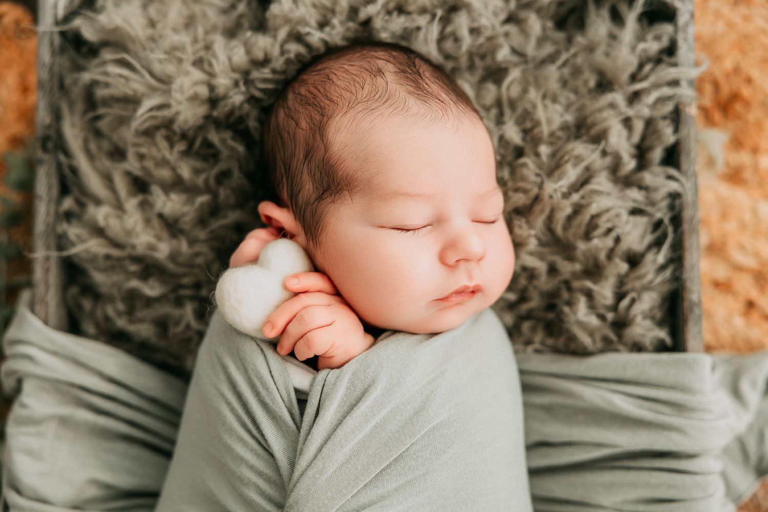 Sleeping newborn baby swaddled in sage green wrap on fluffy gray blanket with white cotton flower prop.