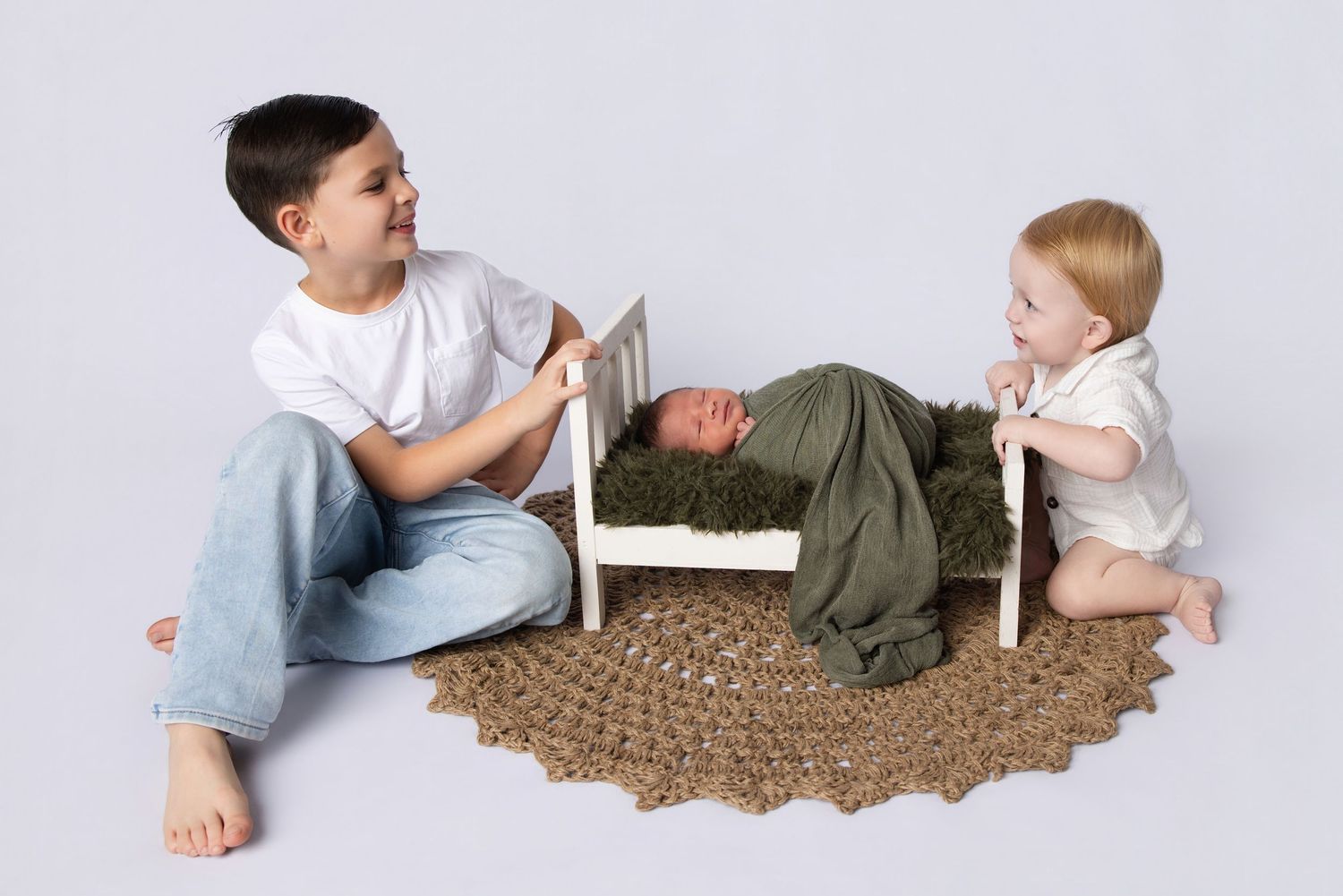 Two older siblings sit beside a tiny newborn resting in a small white bed on a woven rug during a studio photo session.