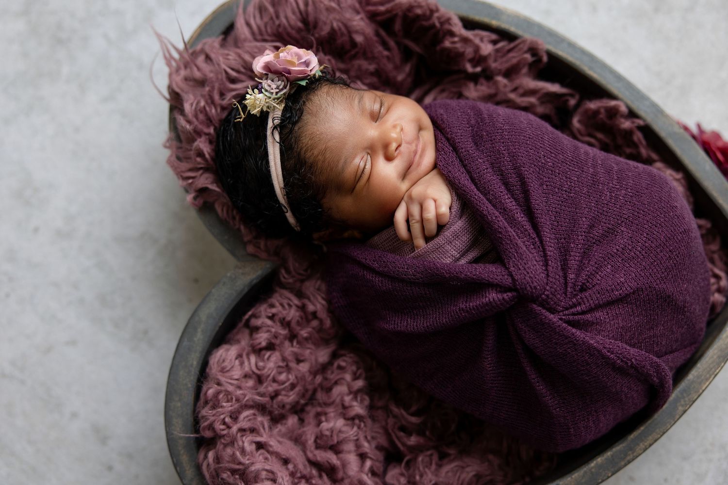 Newborn baby girl swaddled in purple wrap, sleeping peacefully in heart-shaped bowl with floral headband.
