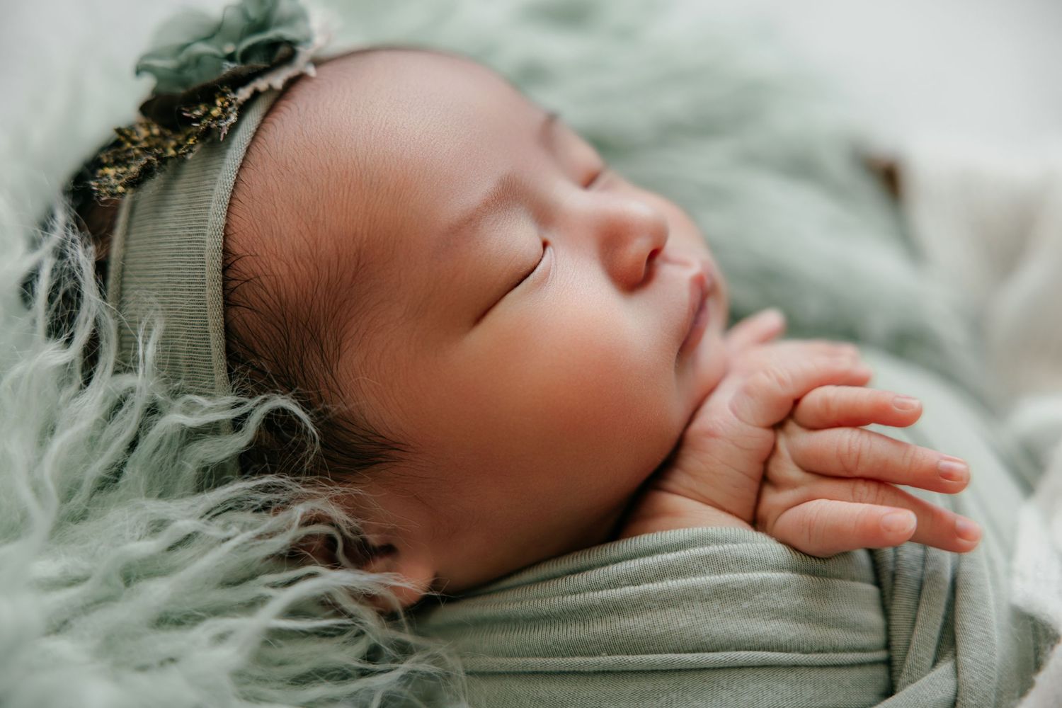 Sleeping newborn baby wrapped in soft green blanket wearing floral headband, peaceful close-up portrait photography.
