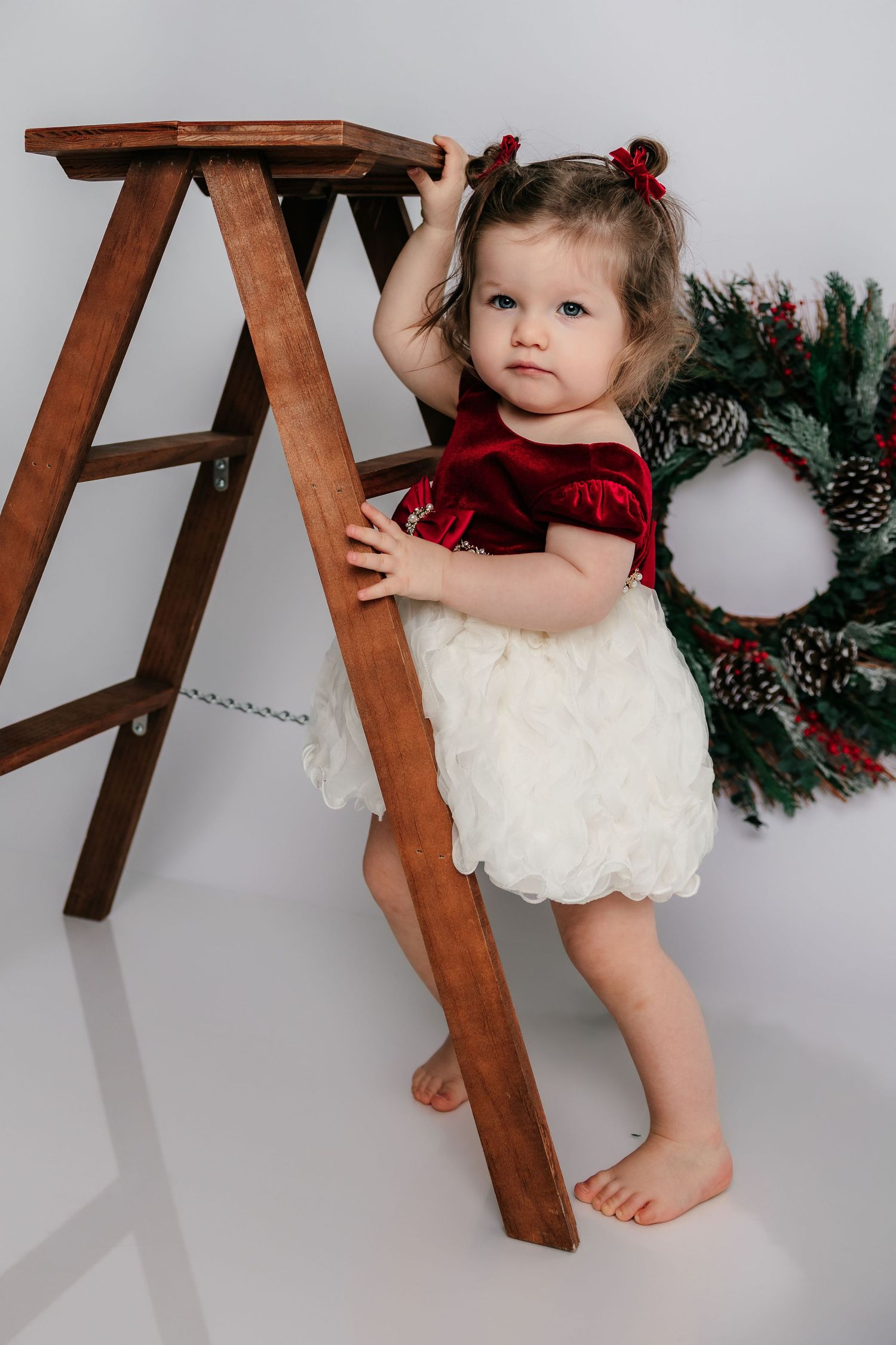 Toddler girl in red velvet and white dress holding wooden ladder near Christmas wreath holiday photo.