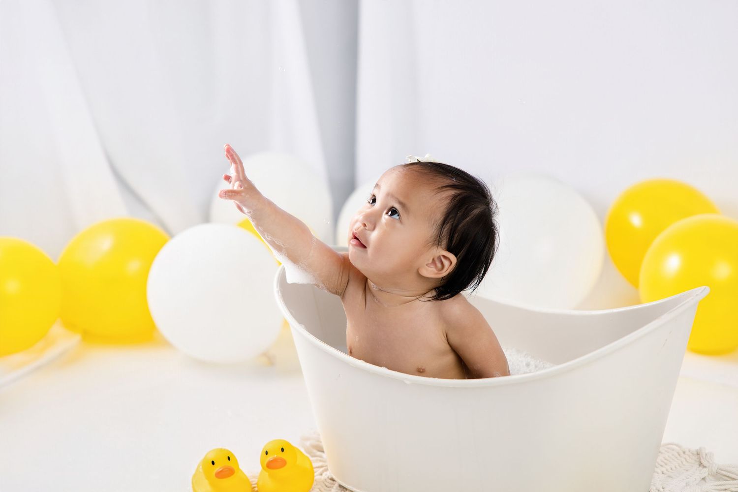 Baby sitting in white tub reaching up, surrounded by yellow rubber ducks and balloons in bright white room.