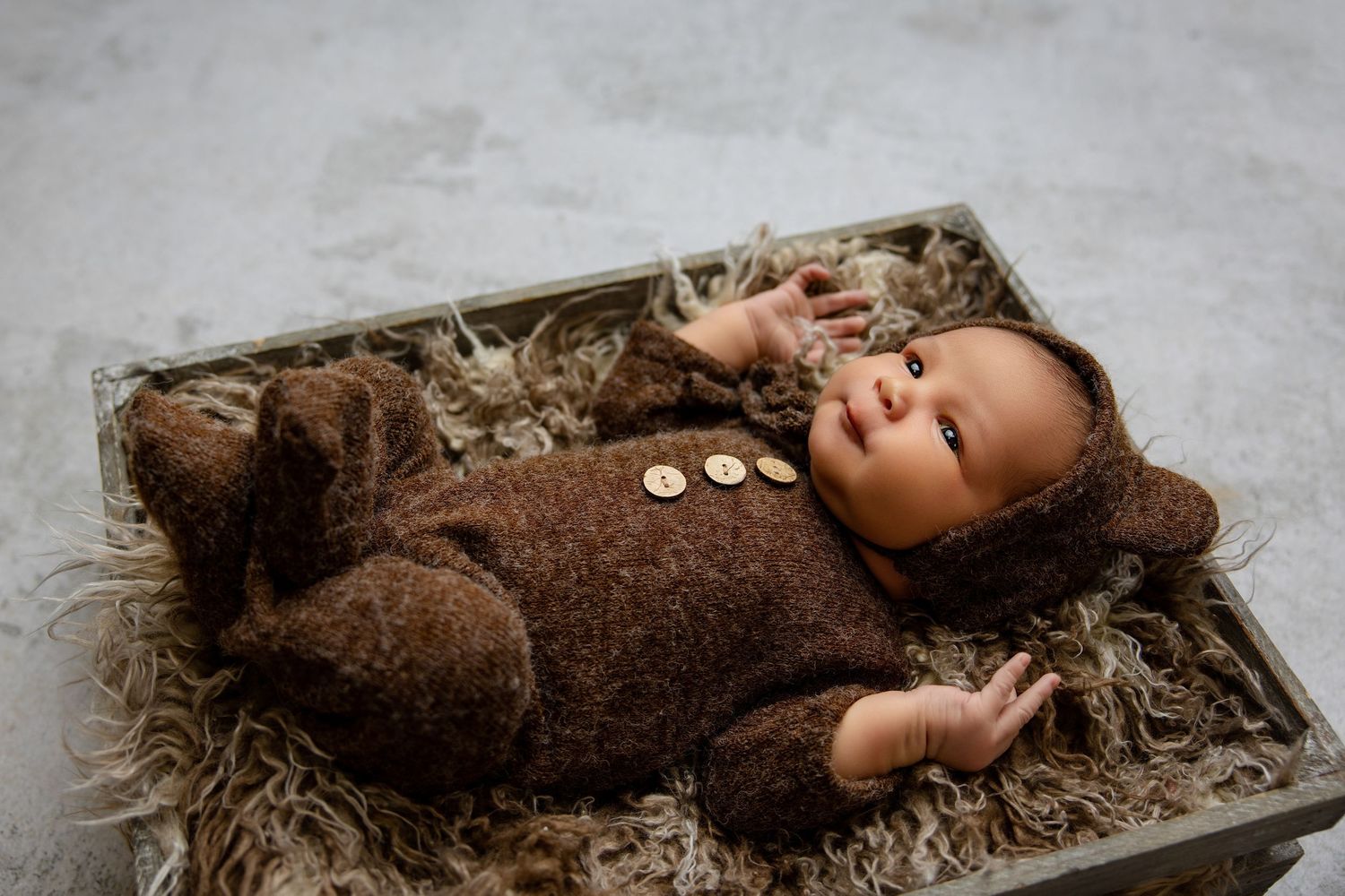 Newborn baby in brown bear knit romper lying in wooden box with fluffy bedding.