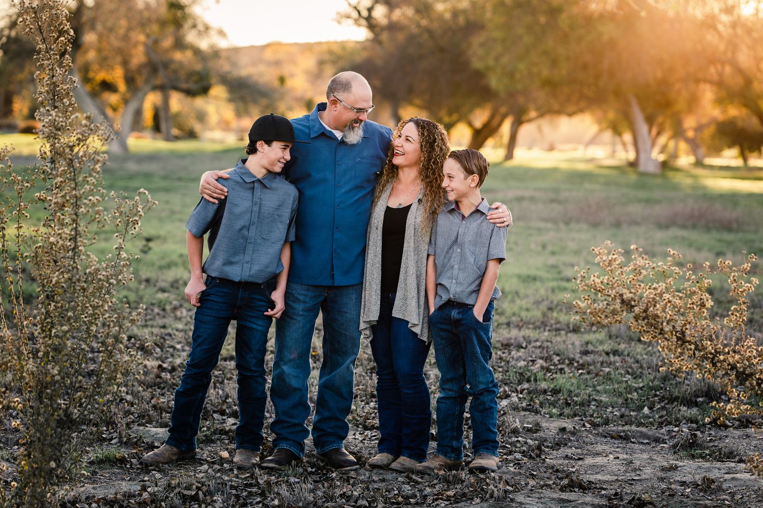 Family of four shares a warm embrace in a golden sunlit field during outdoor portrait.