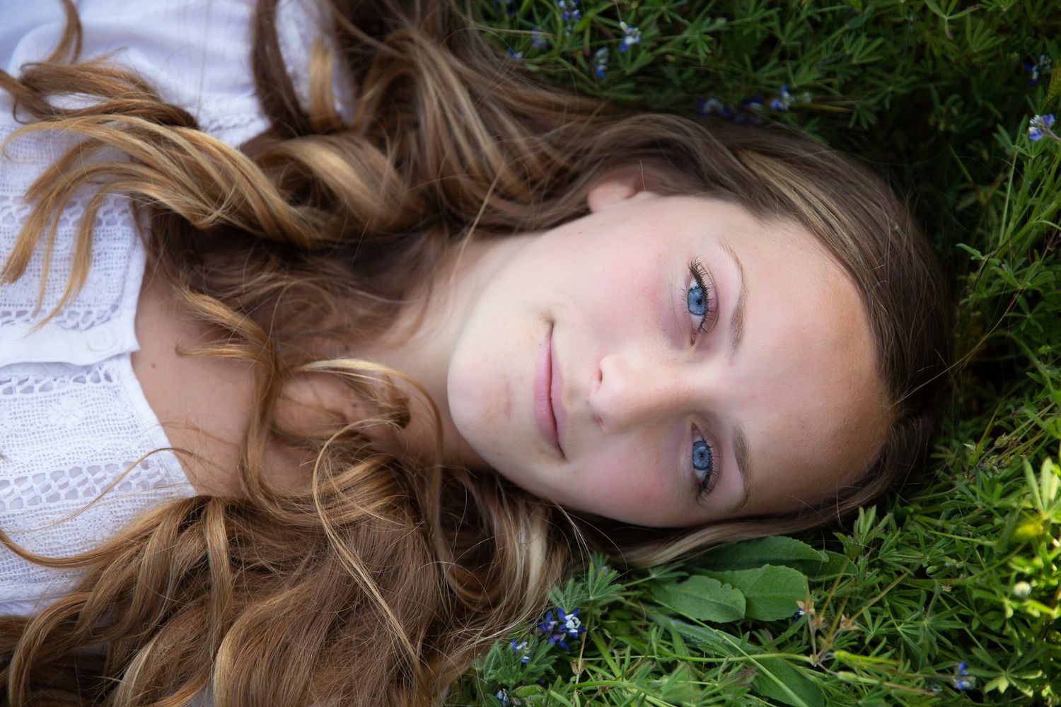 Young girl with blue eyes and wavy hair lies peacefully in green grass with wildflowers. Senior photography session in Vista