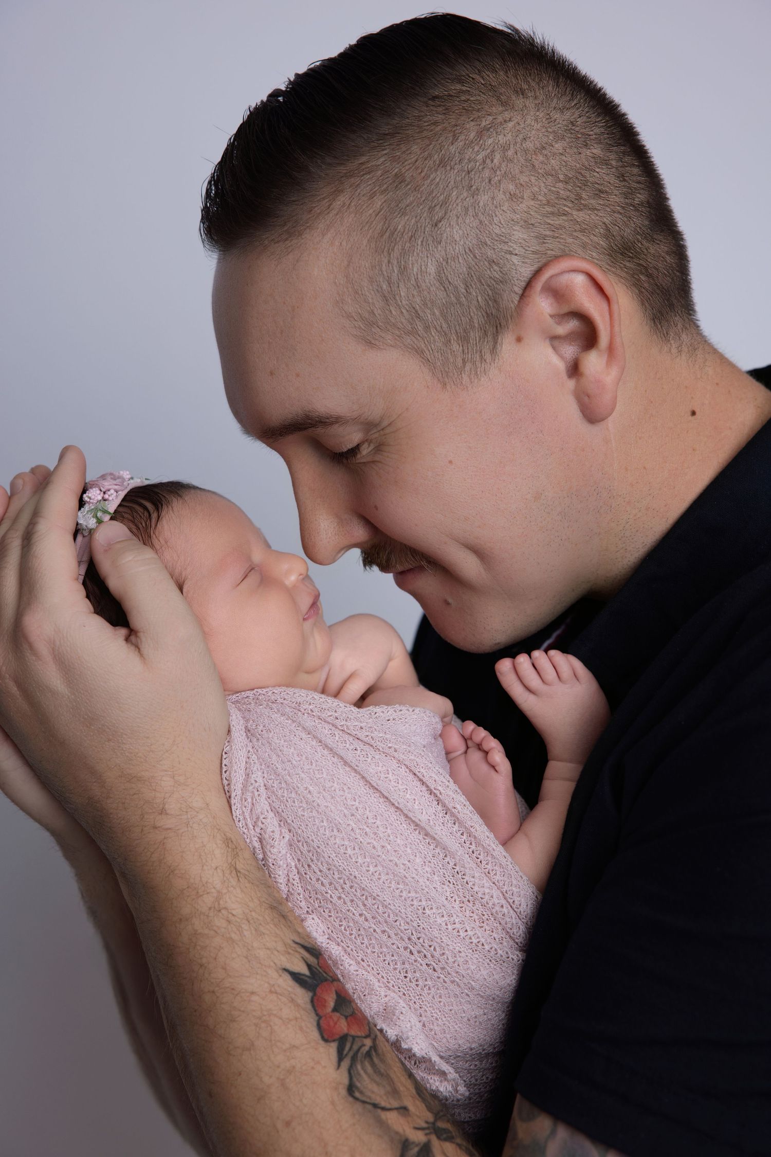 A tattooed father tenderly touches noses with his swaddled newborn baby girl wearing a pink floral headband.