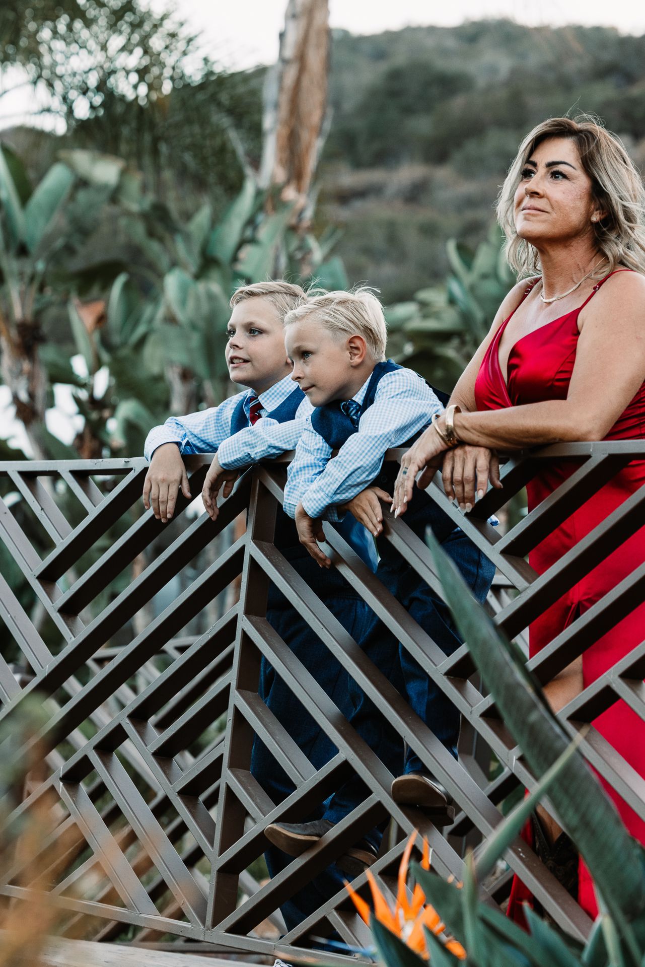 Woman in red dress with two blonde boys in suits leaning on decorative railing.