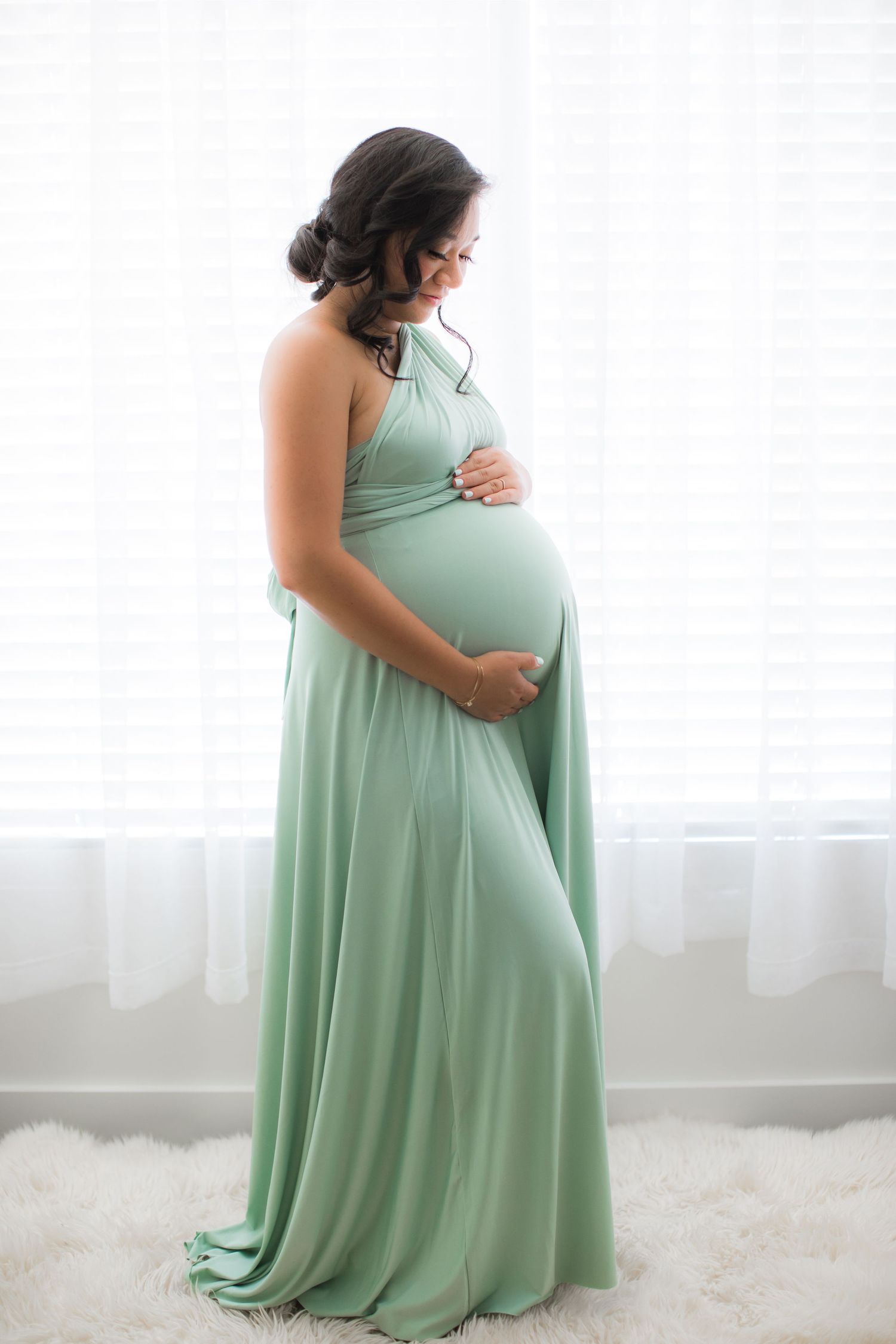 Pregnant woman in a flowing mint green gown tenderly holding her baby bump by a bright window in Bonsall, near Fallbrook, & Temecula