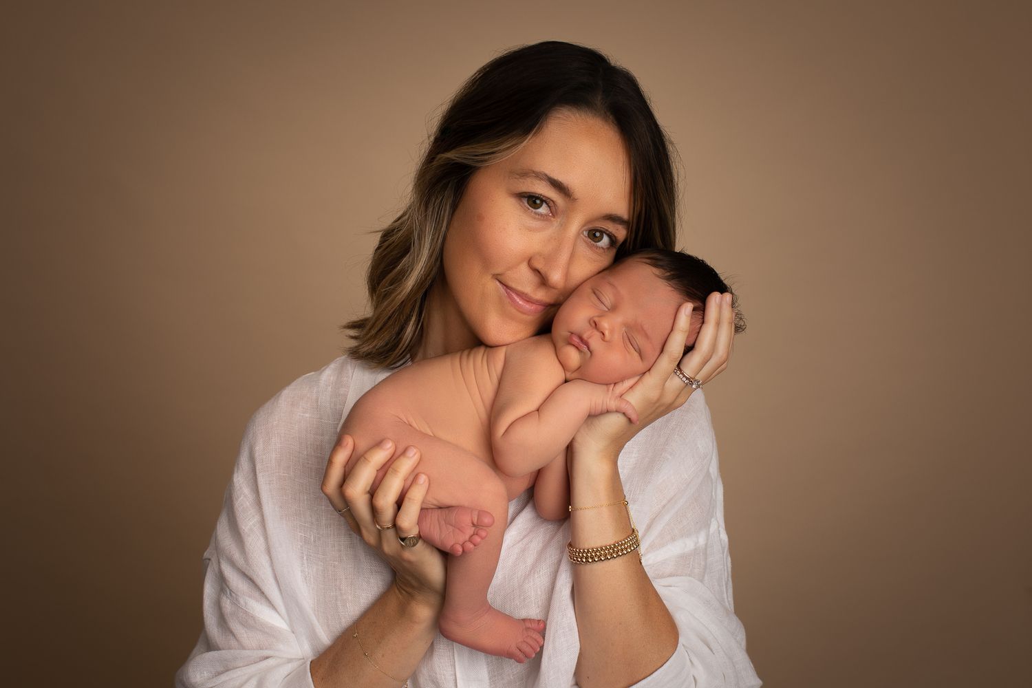 Smiling mother in white shirt holding sleeping newborn baby against beige background.