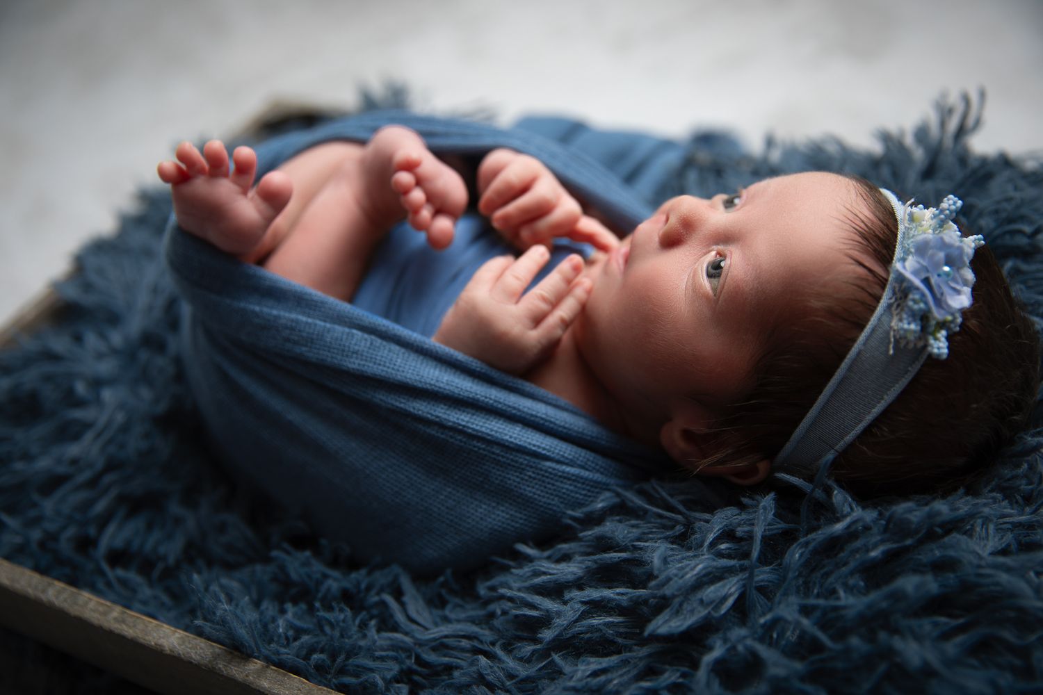 Awake newborn baby wrapped in blue swaddle with floral headband lying on navy blue fluffy fur at The Studio Bonsall, near Fallbrook