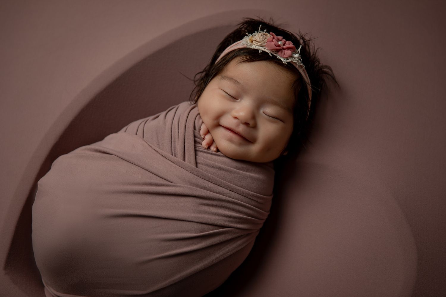 Smiling newborn baby swaddled in mauve wrap wearing a colorful floral headband against a pink background.