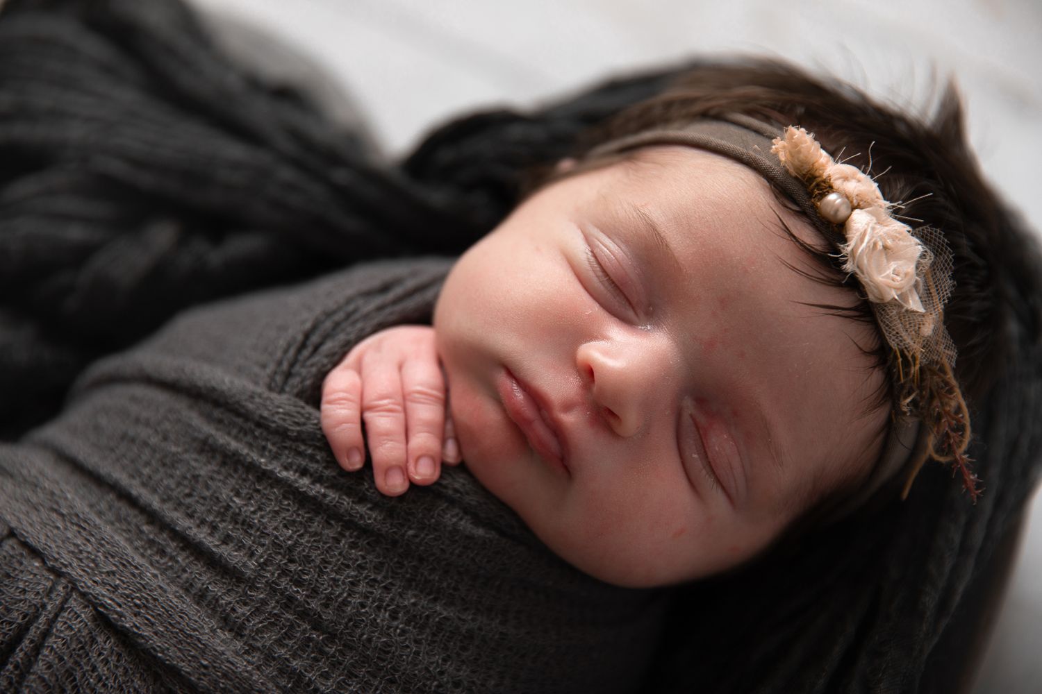 Sleeping newborn baby swaddled in dark gray wrap wearing a delicate floral headband on white background at The Studio Bonsall, near Fallbroo