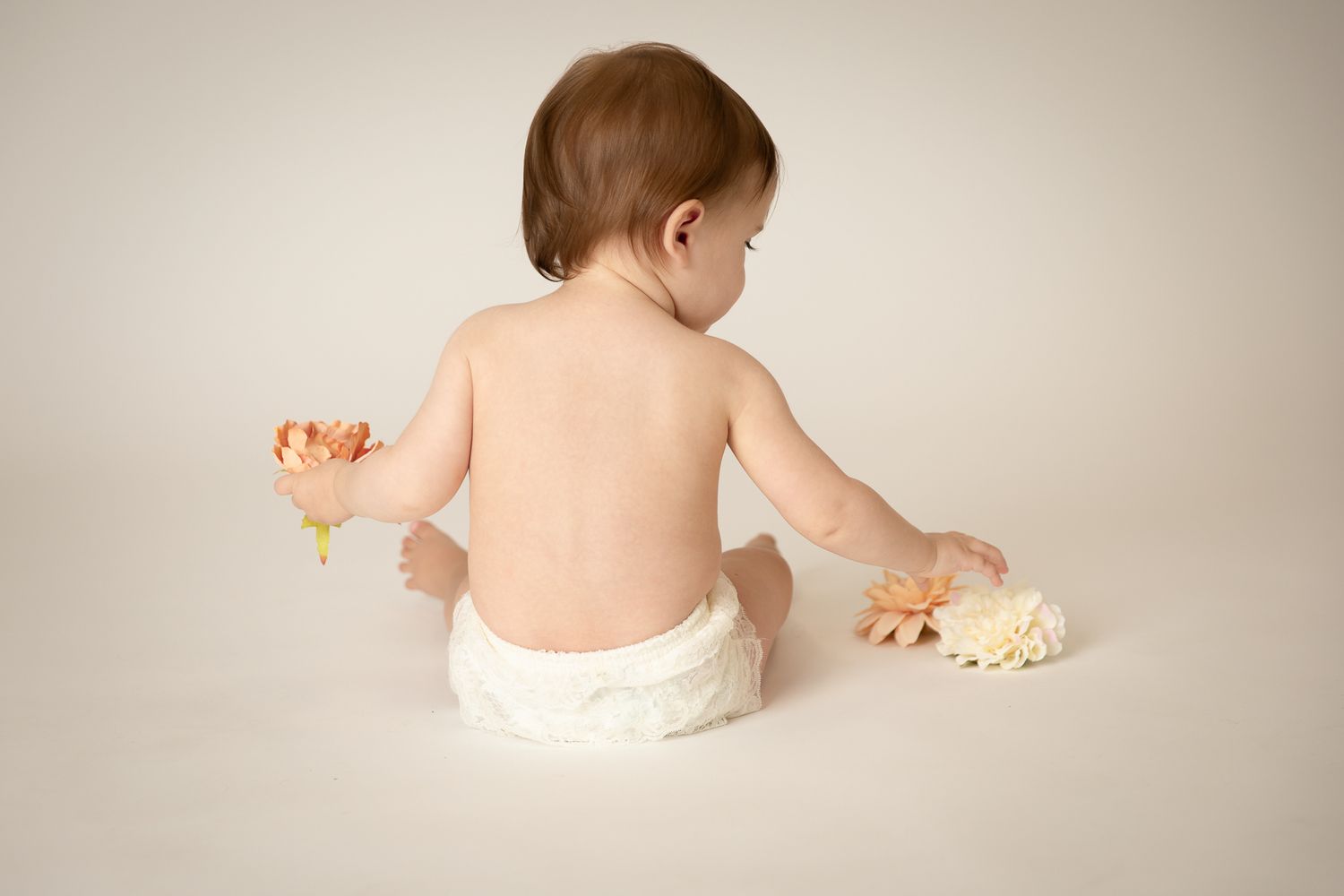 Baby in white diaper sitting with flowers, photographed from behind on a cream background.
